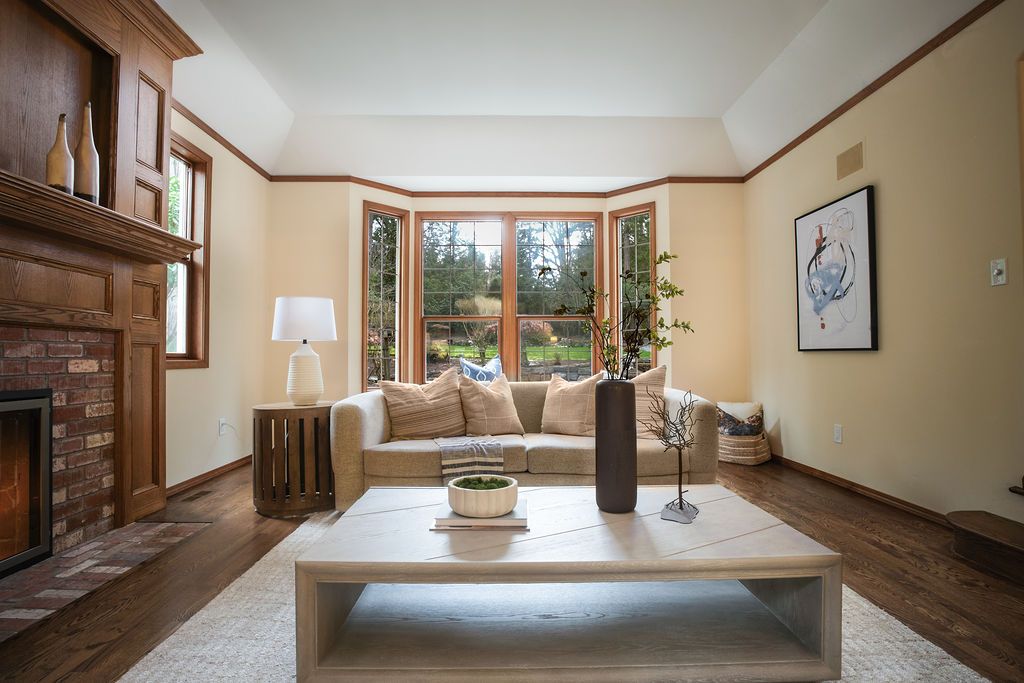 Bright living room with bay windows, brick fireplace, cream sofa, wood coffee table, and hardwood floors.