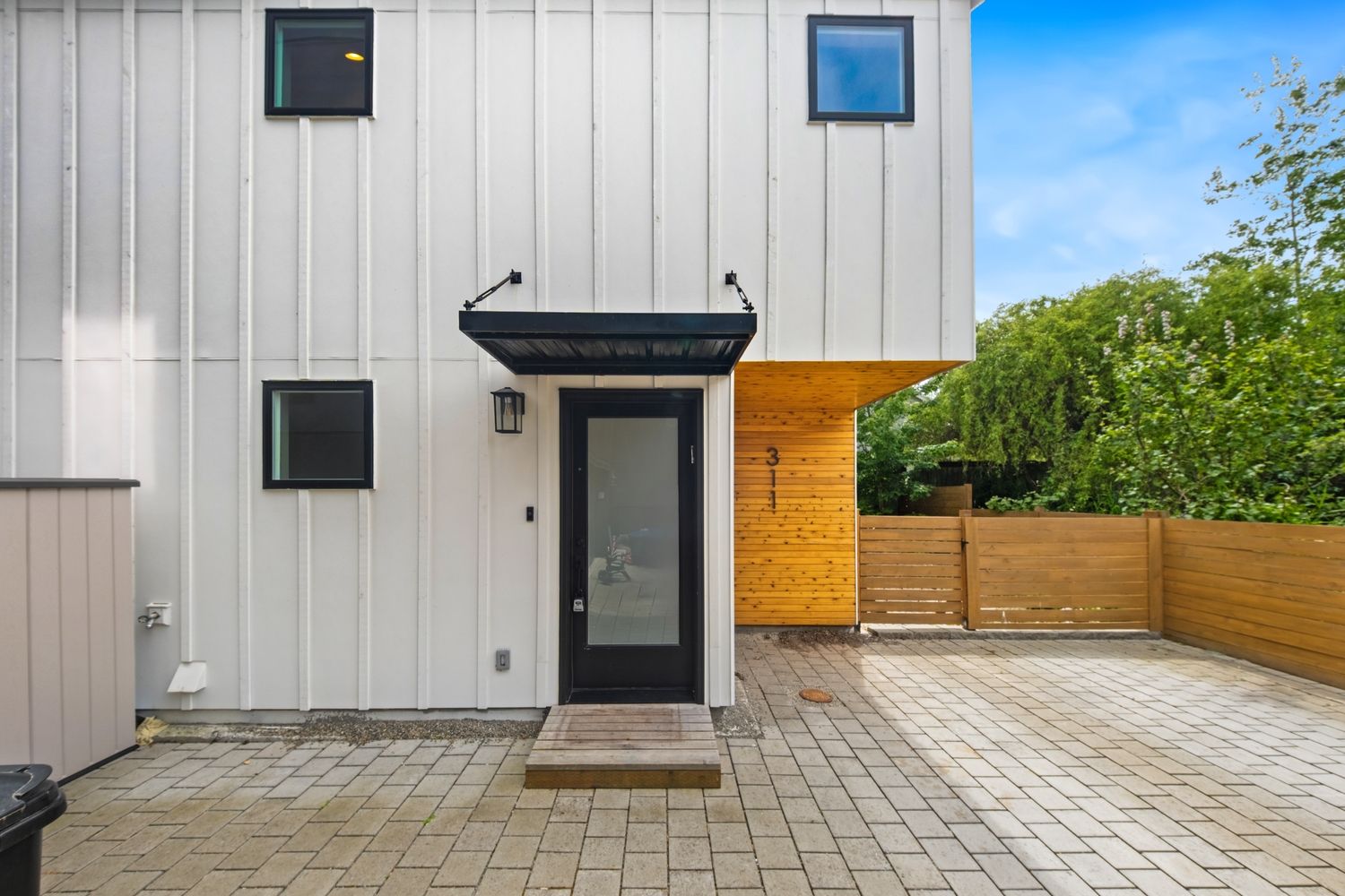 Modern white vertical siding townhouse exterior with black door, covered entry, brick paver patio, and wood fence.