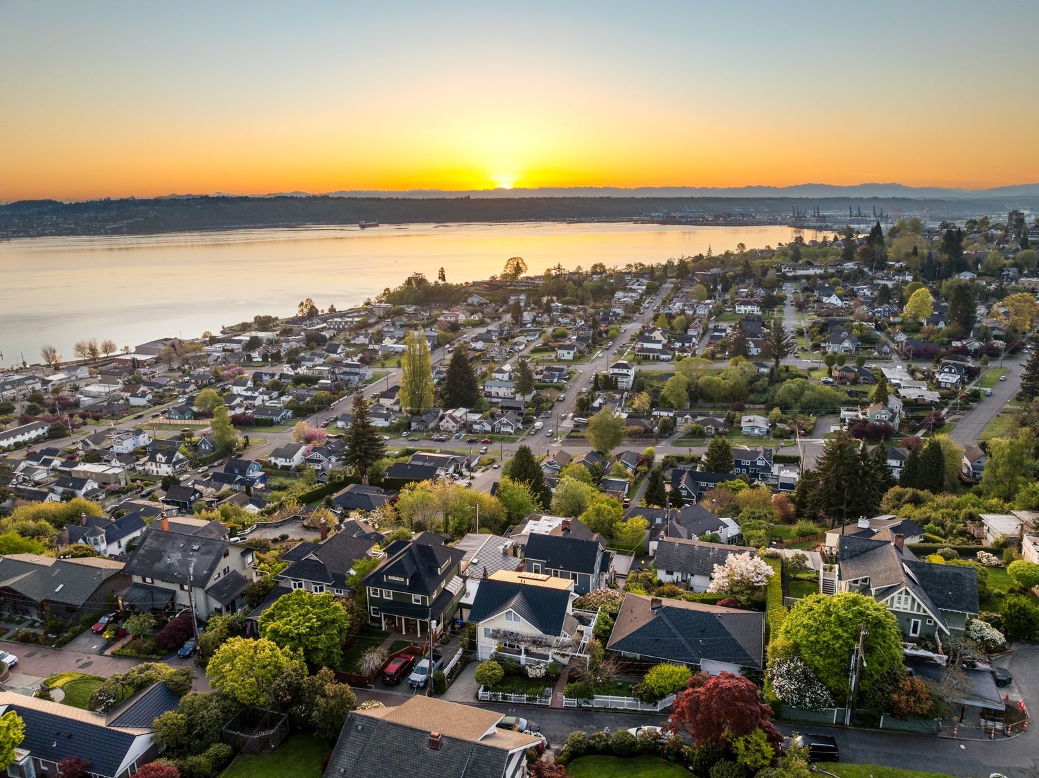 Aerial sunset view over a coastal residential neighborhood with tree-lined streets and waterfront in the background.