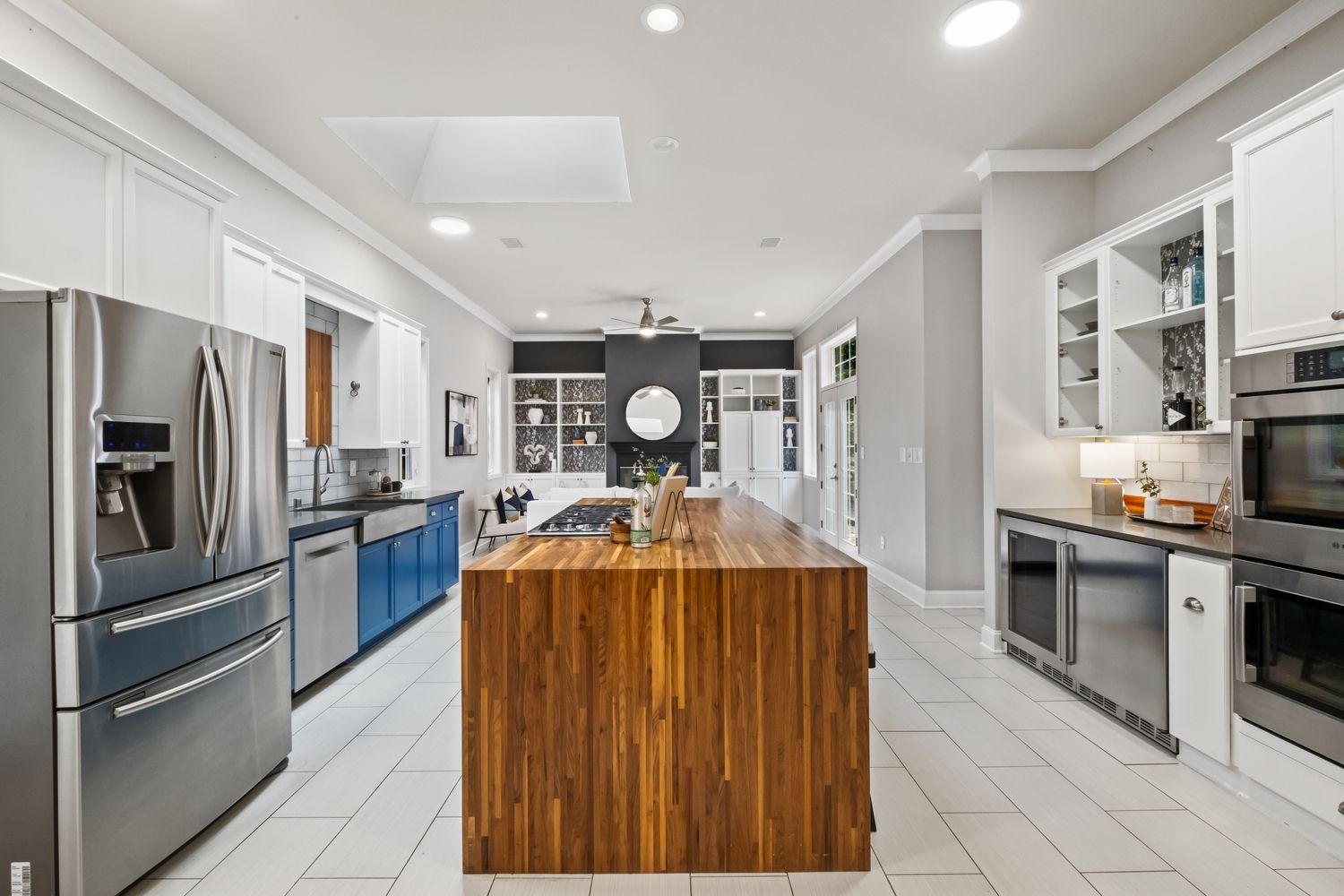 Modern white kitchen with stainless steel appliances and large wooden island featuring bright recessed lighting and tile flooring.