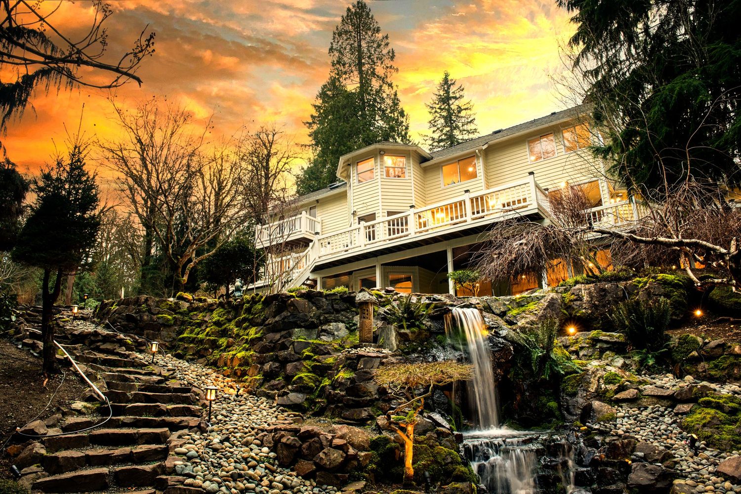 Stunning multi-level home with waterfall, stone steps, lush greenery, and dramatic orange sunset sky.