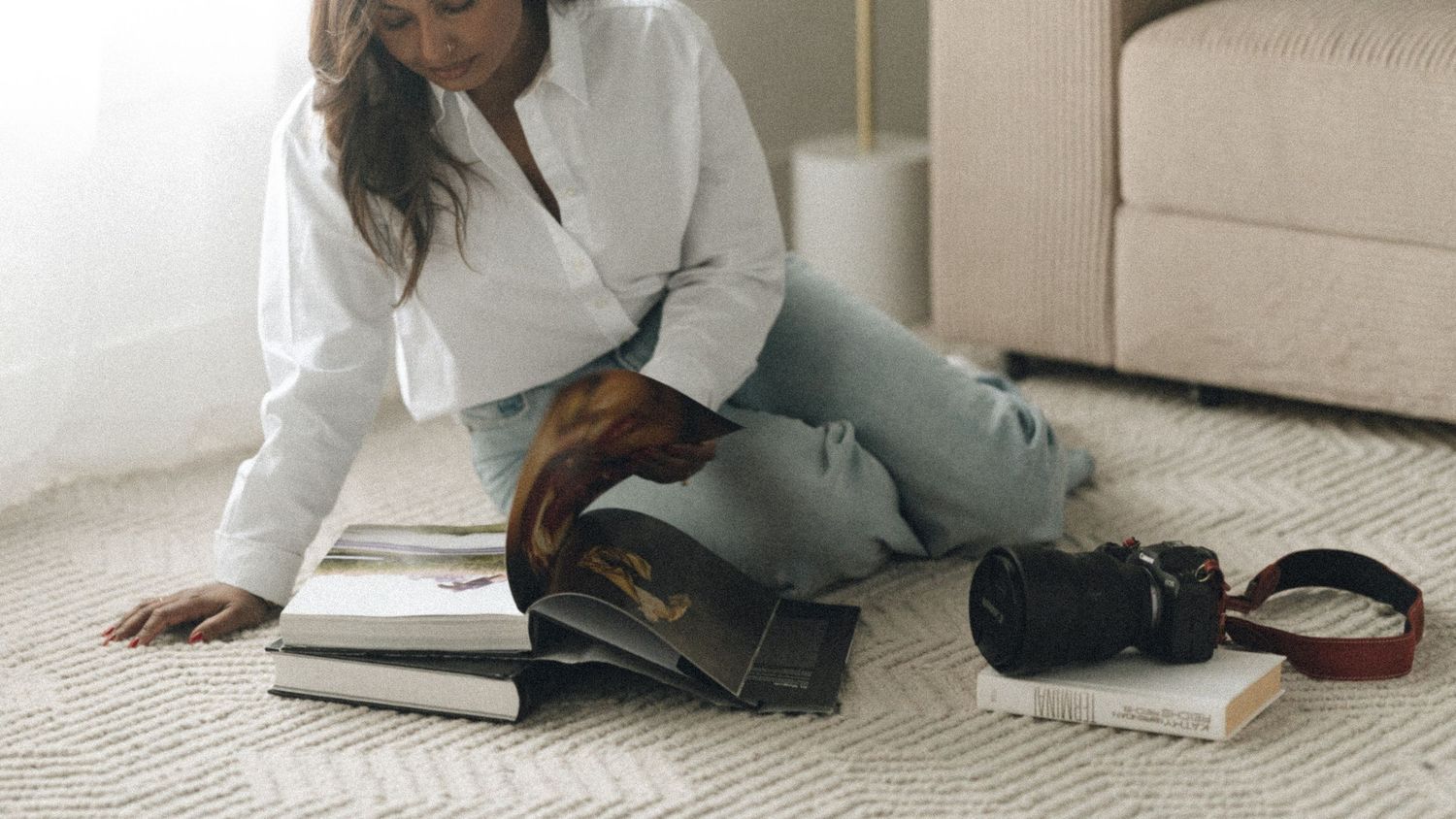 Person in a white shirt sits on carpet reading books with a backpack and other items scattered nearby.
