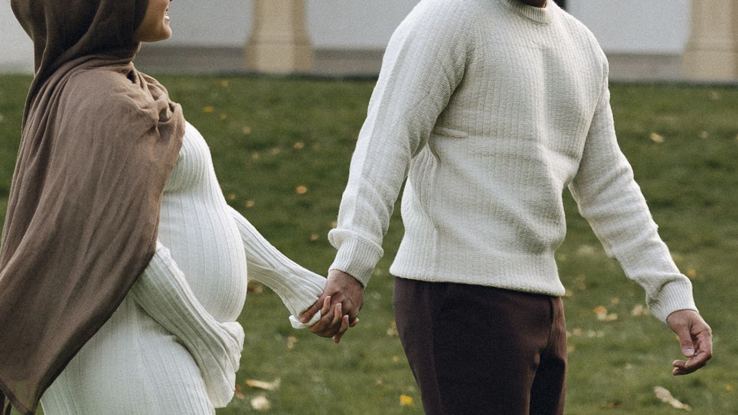 A couple holds hands while walking on grass, with one wearing a white sweater and the other in a brown cardigan.