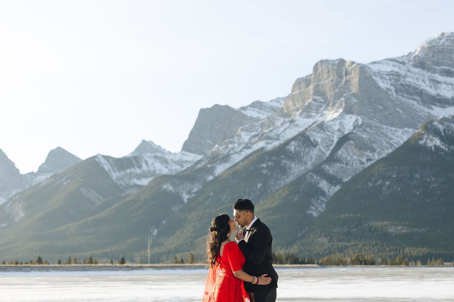A couple in formal wear embraces on a frozen lake with snow-capped Rocky Mountains towering in the background.