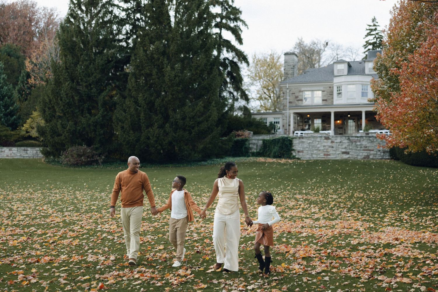A family walks together across a fallen leaf-covered lawn towards a stately white house on an autumn day.