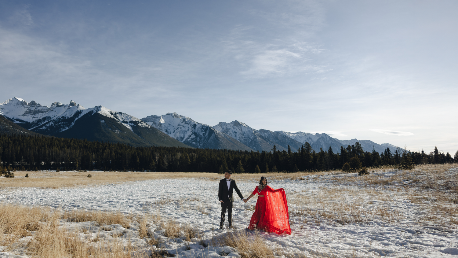 A couple in winter attire holds hands in a snowy field with majestic mountain peaks and blue skies in the background.