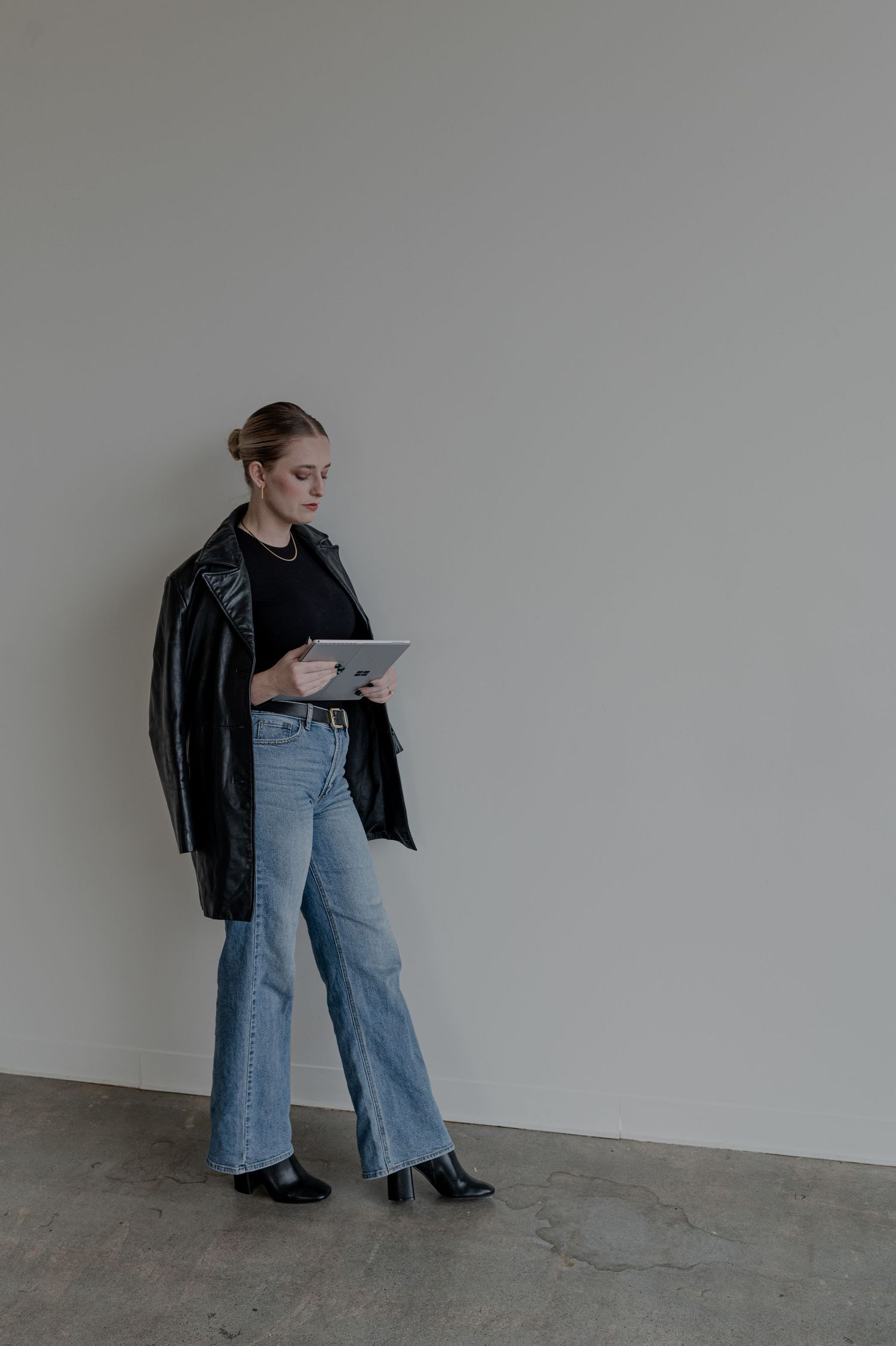 Person wearing leather jacket and jeans standing against grey wall while looking at tablet.