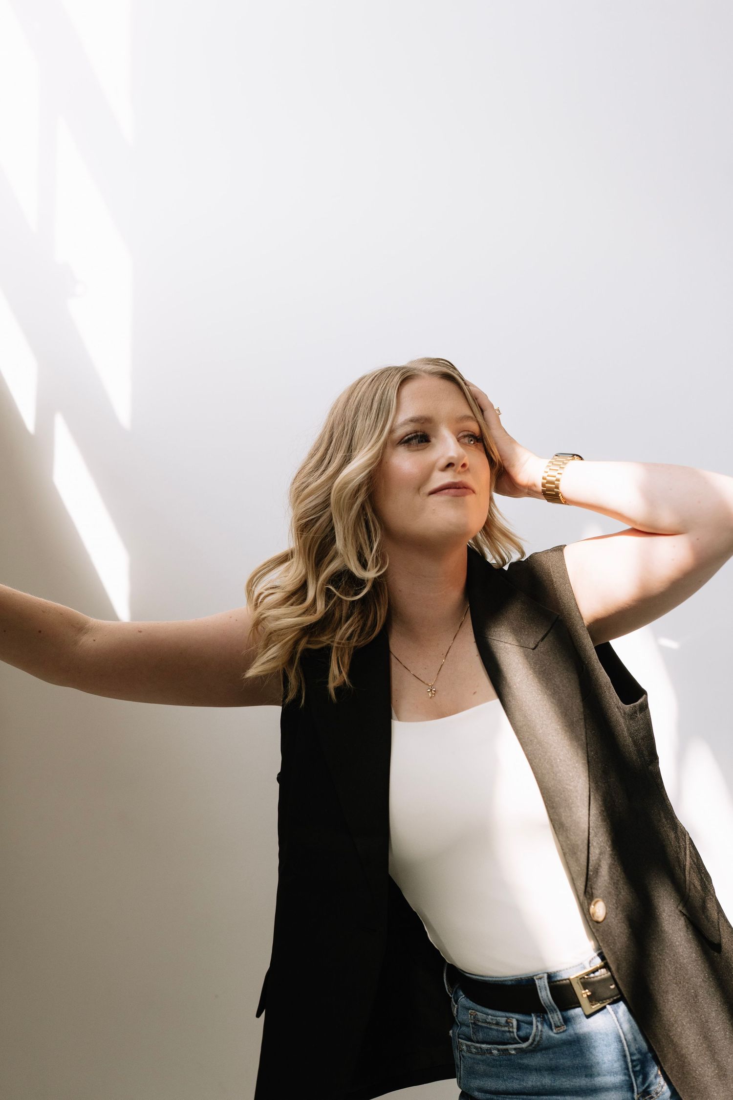 Blonde woman in black vest, white top, and jeans poses confidently against a white wall in natural light.