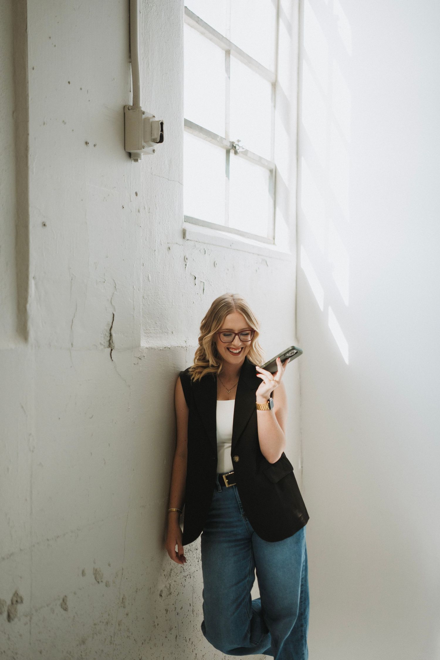 Smiling blonde woman in glasses and black vest leans against white wall holding a phone in bright studio space.