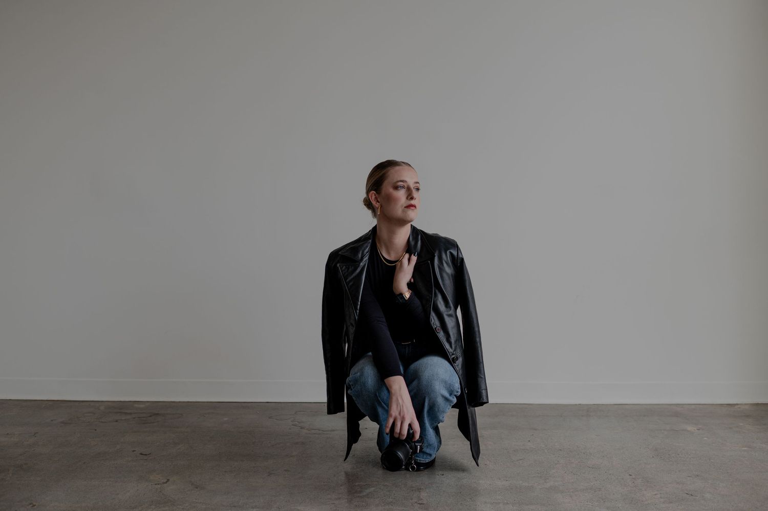 Person in leather jacket and jeans crouching against minimal grey wall.