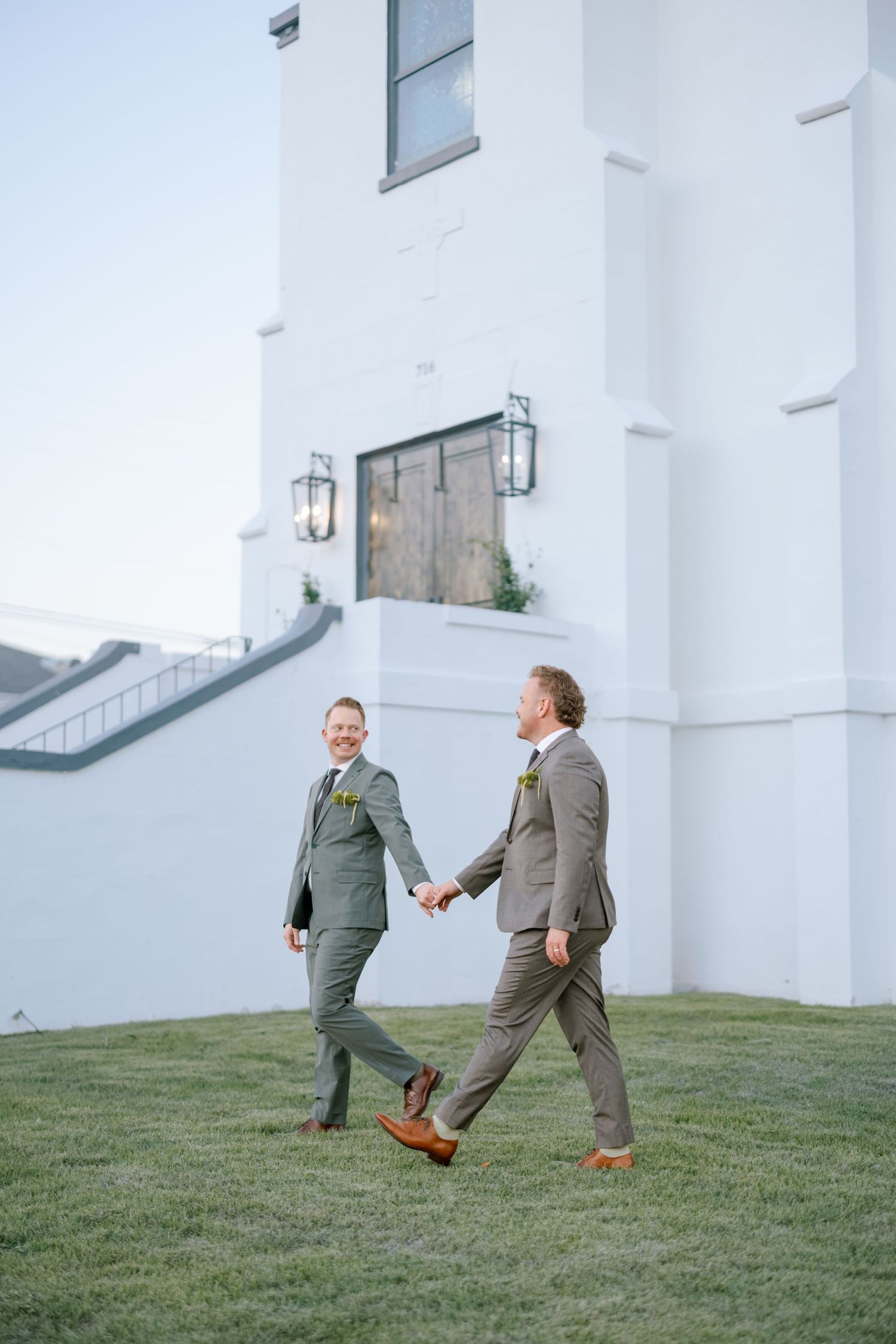 Two grooms in suits playfully chase each other on a grassy hill outside a white building.