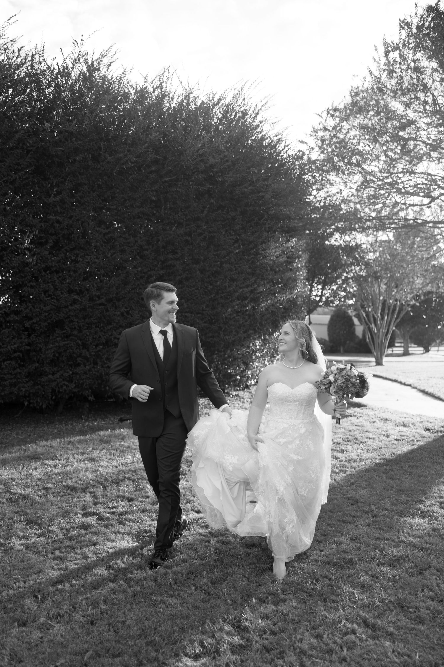 A bride in a flowing white dress and groom in dark suit walk together on a grassy path beside hedges.