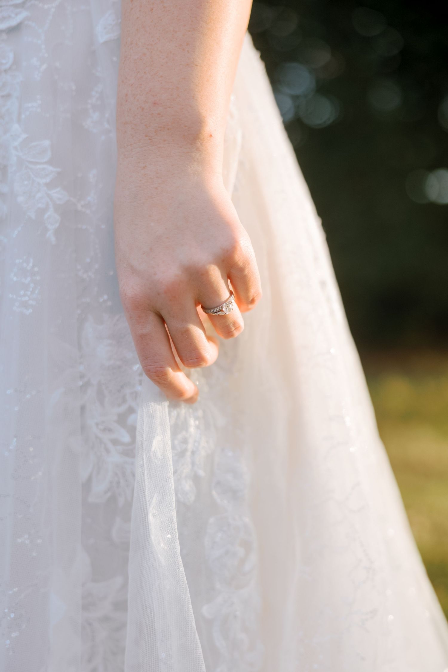 Close-up detail of a white lace wedding dress being held delicately by a hand wearing an engagement ring.