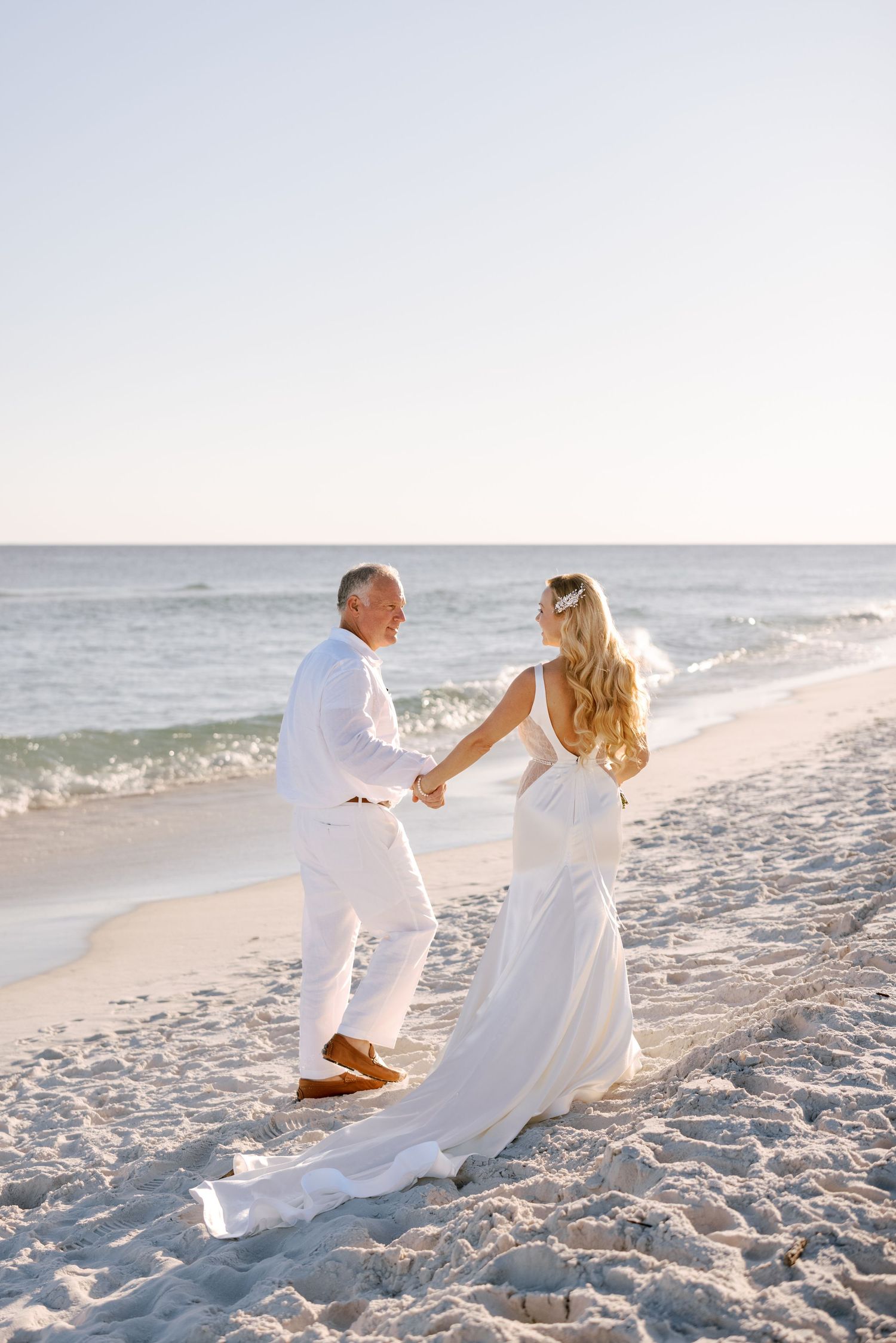 A couple in white outfits dance together on a sunny beach with waves in the background.