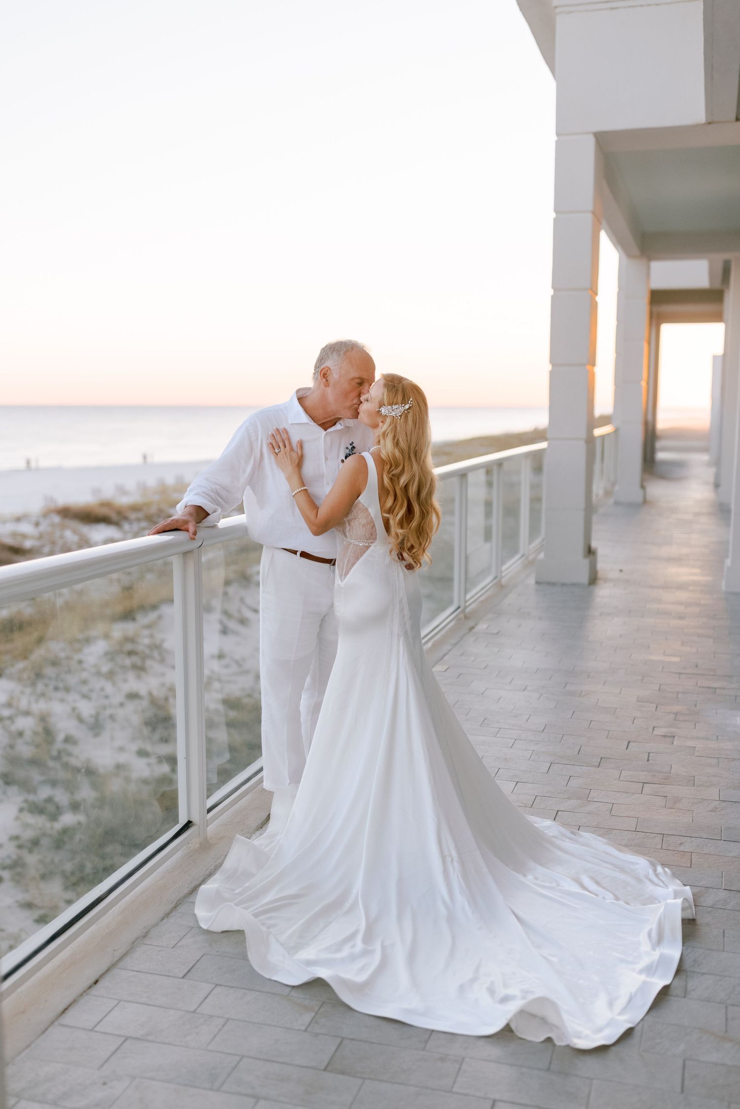 A romantic couple in white attire share an intimate dance moment on a beachfront balcony at sunset.