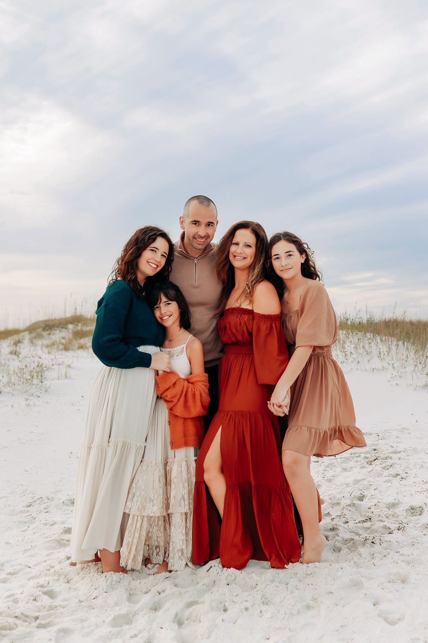 A group in coordinated rust and cream colored dresses poses together on a white sandy beach at sunset in Destin, Florida.