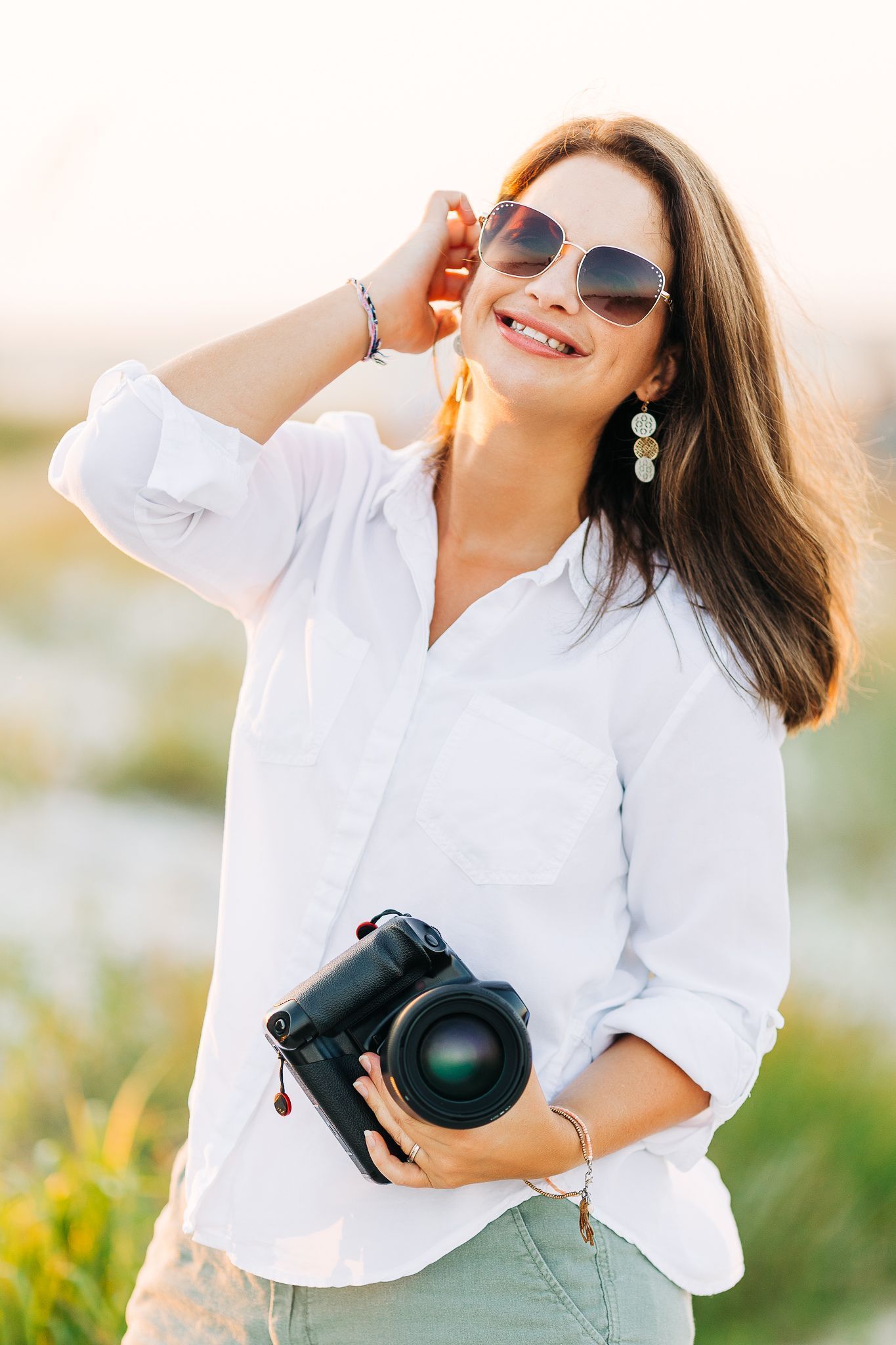 Photographer in white shirt and sunglasses poses with camera in outdoor natural setting during golden hour in Destin, FL.