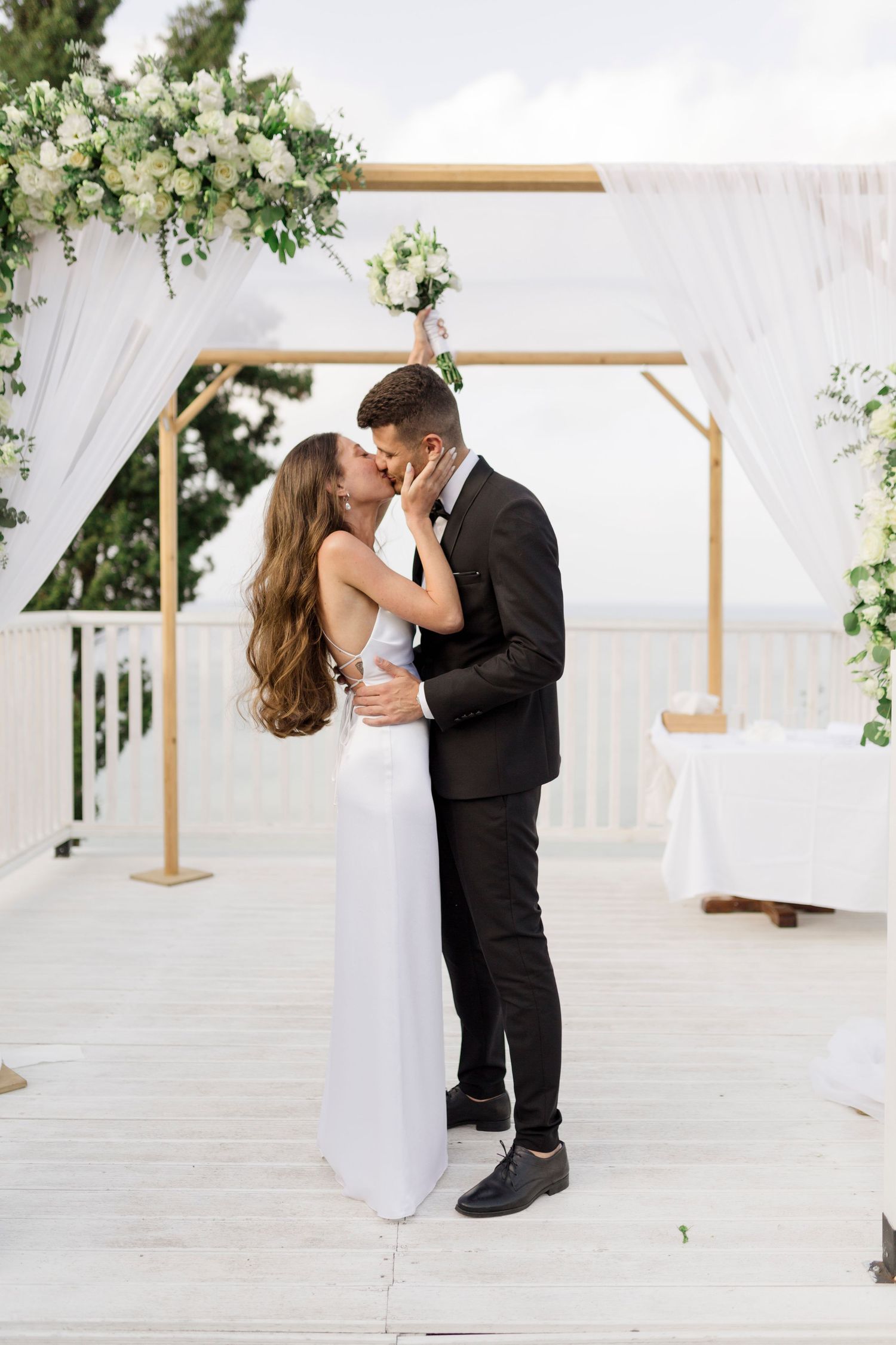 A romantic wedding ceremony under an elegant white draped arch decorated with lush white and green floral arrangements.