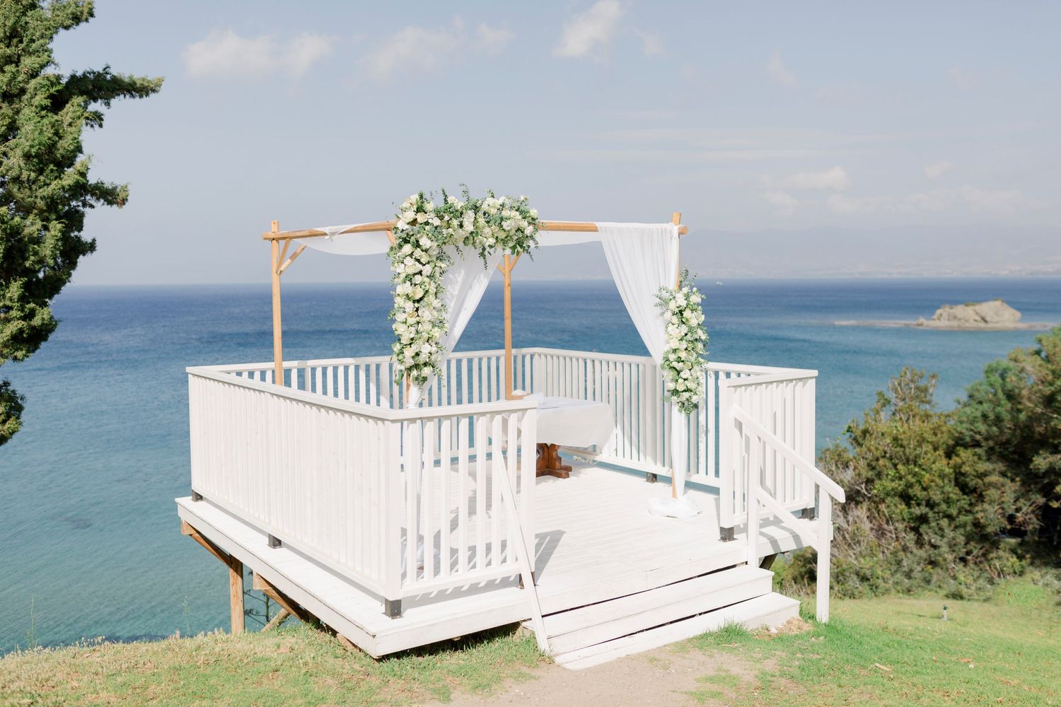 A beautiful white wedding gazebo decorated with floral arrangements overlooks a turquoise ocean on a hilltop.