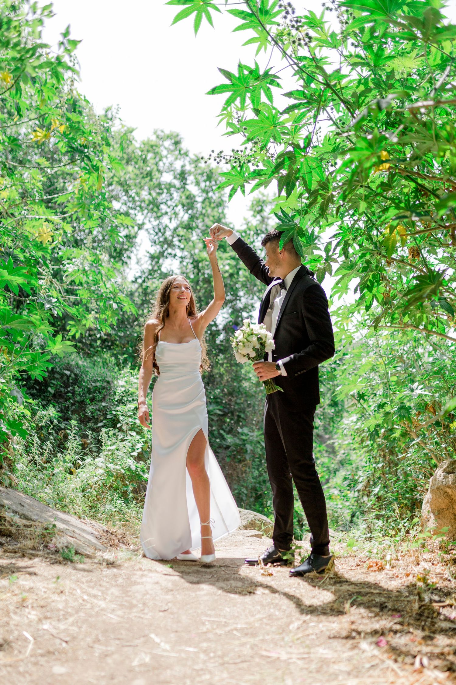 Couple in elegant wedding attire sharing a joyous moment on a lush green garden path.