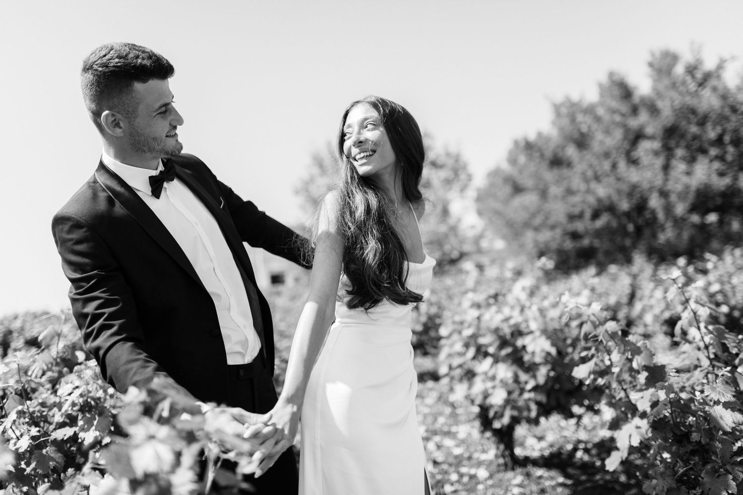 A black and white wedding photo showing a couple holding hands and laughing together in a garden setting.