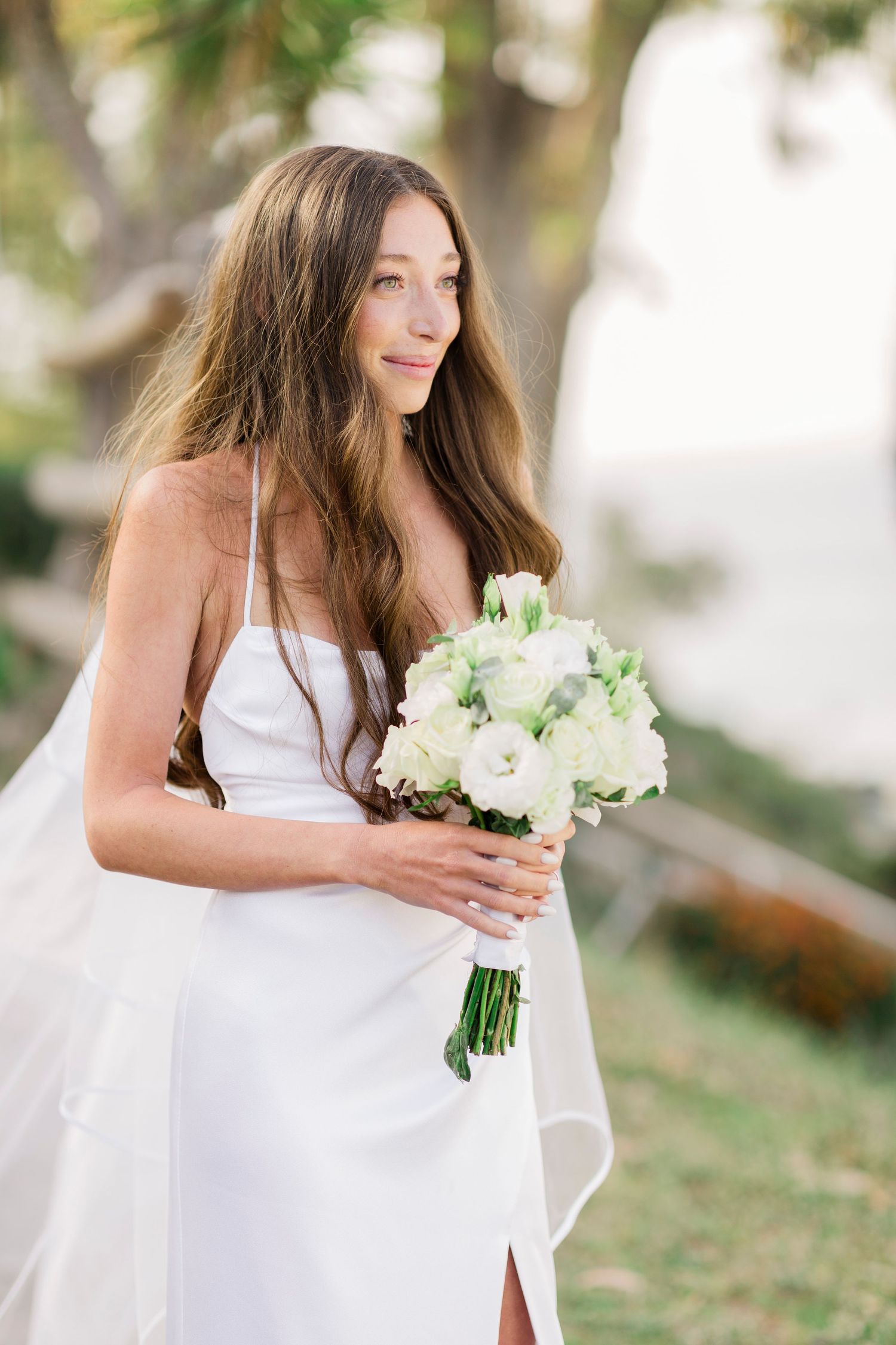A bride in a sleek white dress holding a bouquet of white flowers against a natural backdrop.