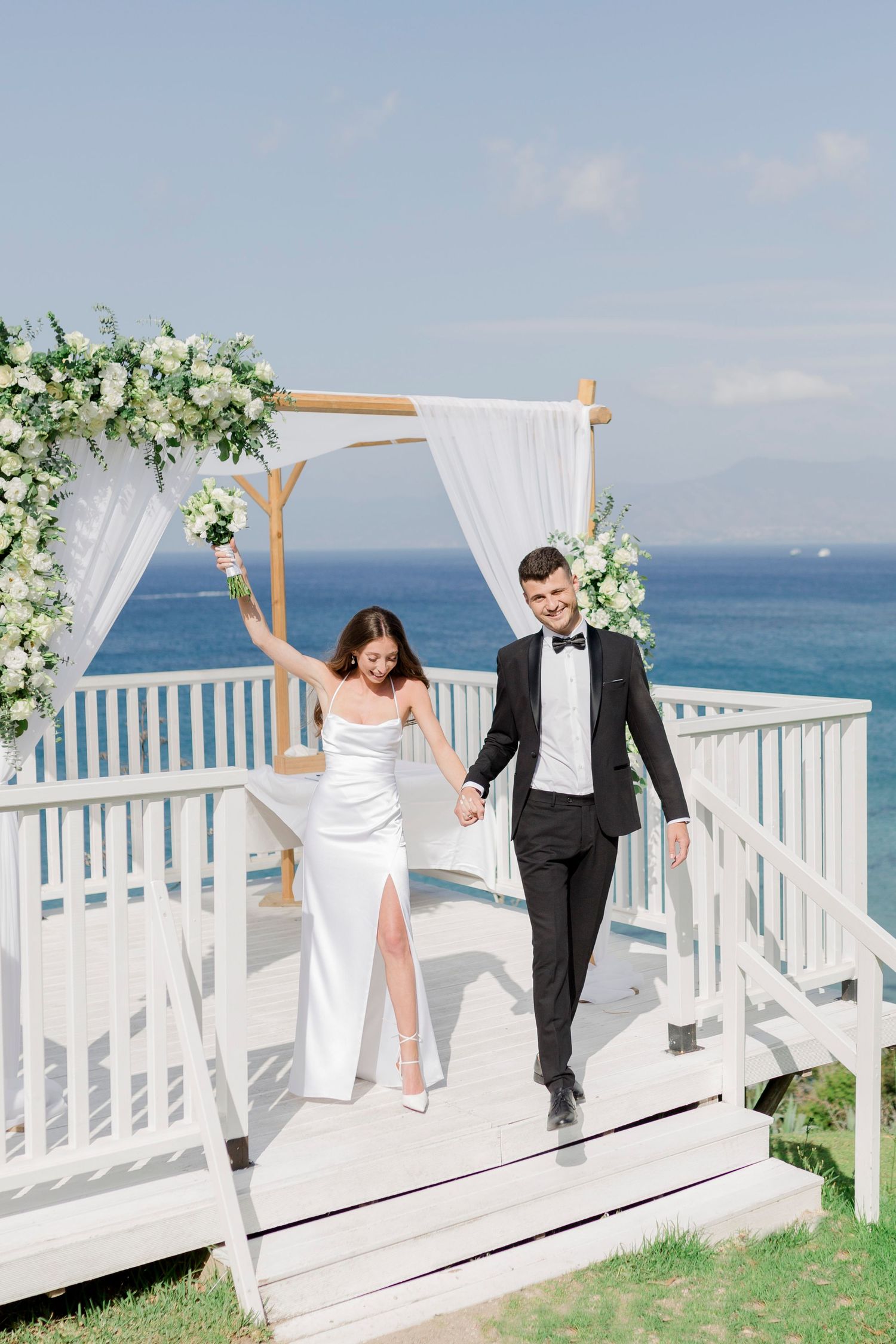 A couple in wedding attire walk together on a coastal white deck with floral arch and ocean backdrop.