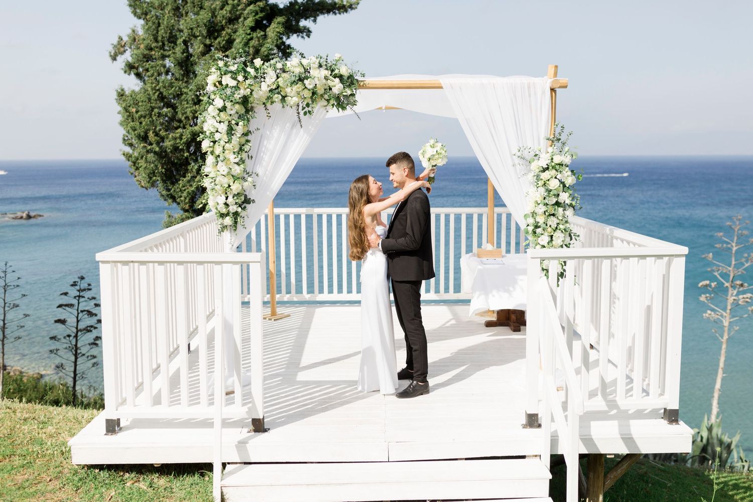 A romantic wedding ceremony takes place on a white gazebo overlooking the ocean with floral decorations.