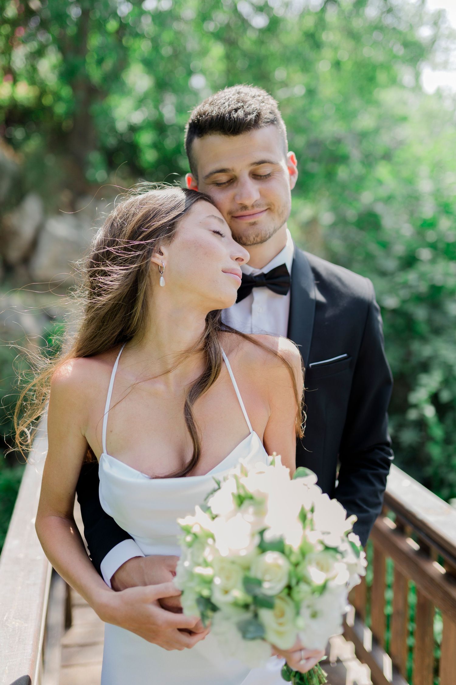 A romantic wedding couple embraces outdoors with the bride holding a white floral bouquet against a green garden backdrop.