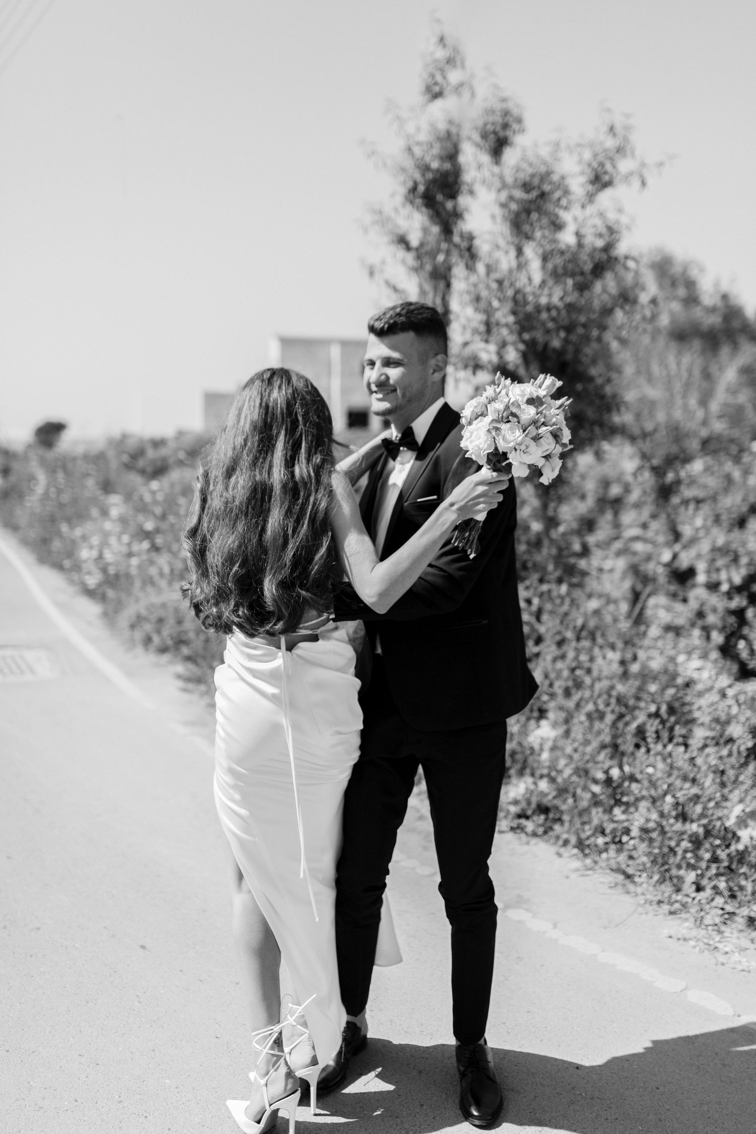 Black and white photo of a couple in wedding attire embracing on a rural path with the bride holding a floral bouquet.