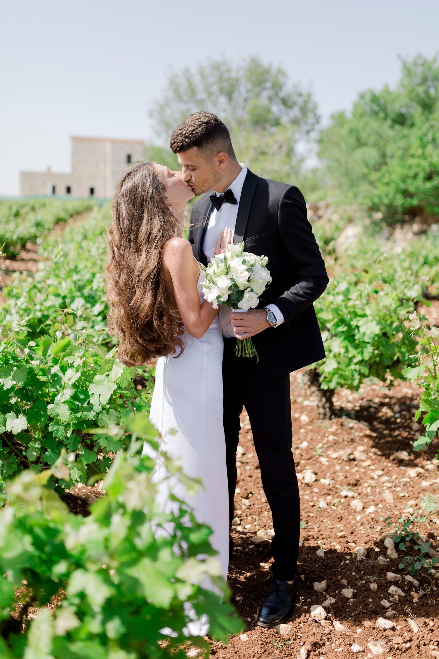 Romantic wedding photoshoot in a lush green vineyard with a couple holding a white bouquet and sharing an intimate moment.