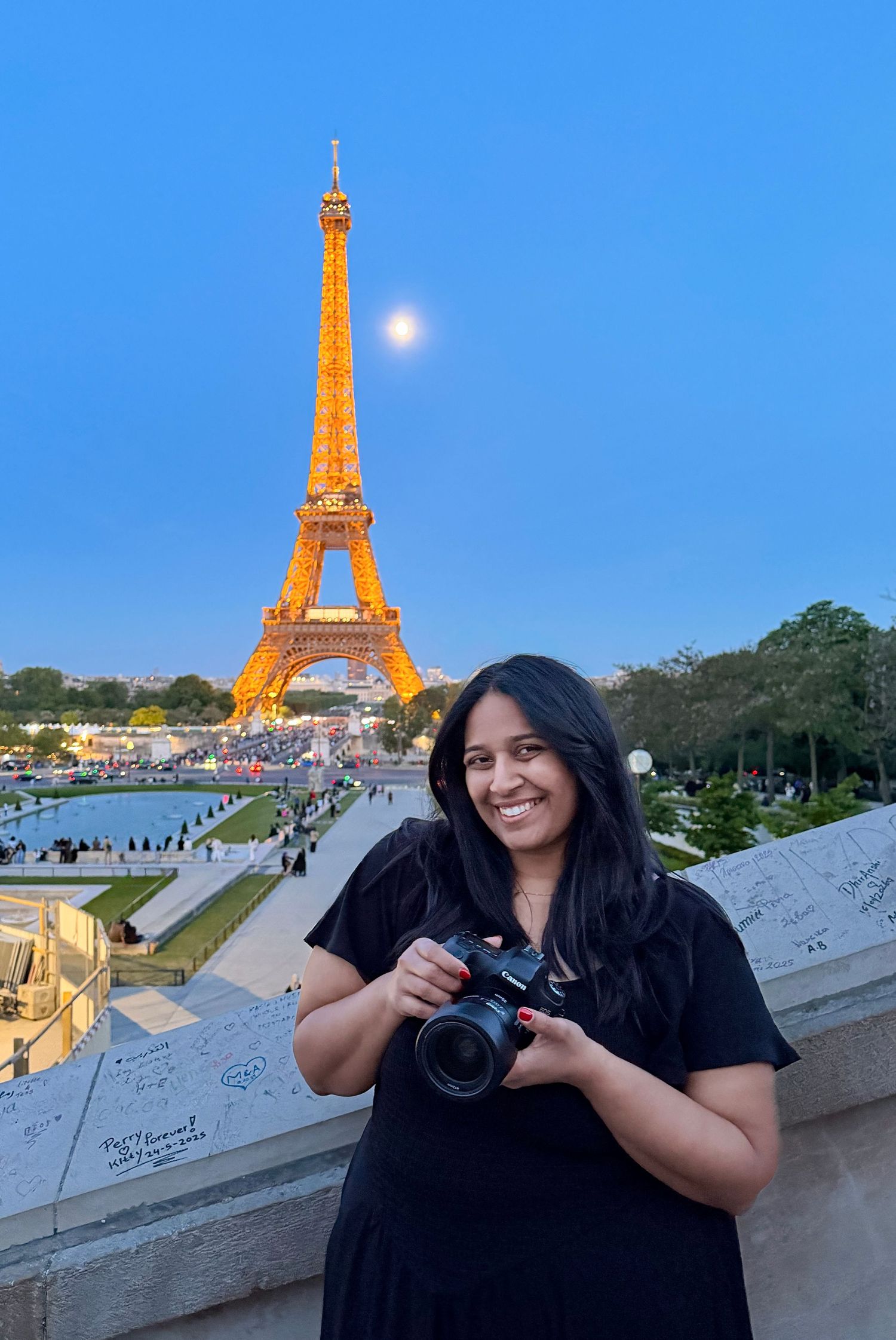 Female wedding photographer smiling with camera in front of the illuminated Eiffel Tower at dusk in Paris, France.