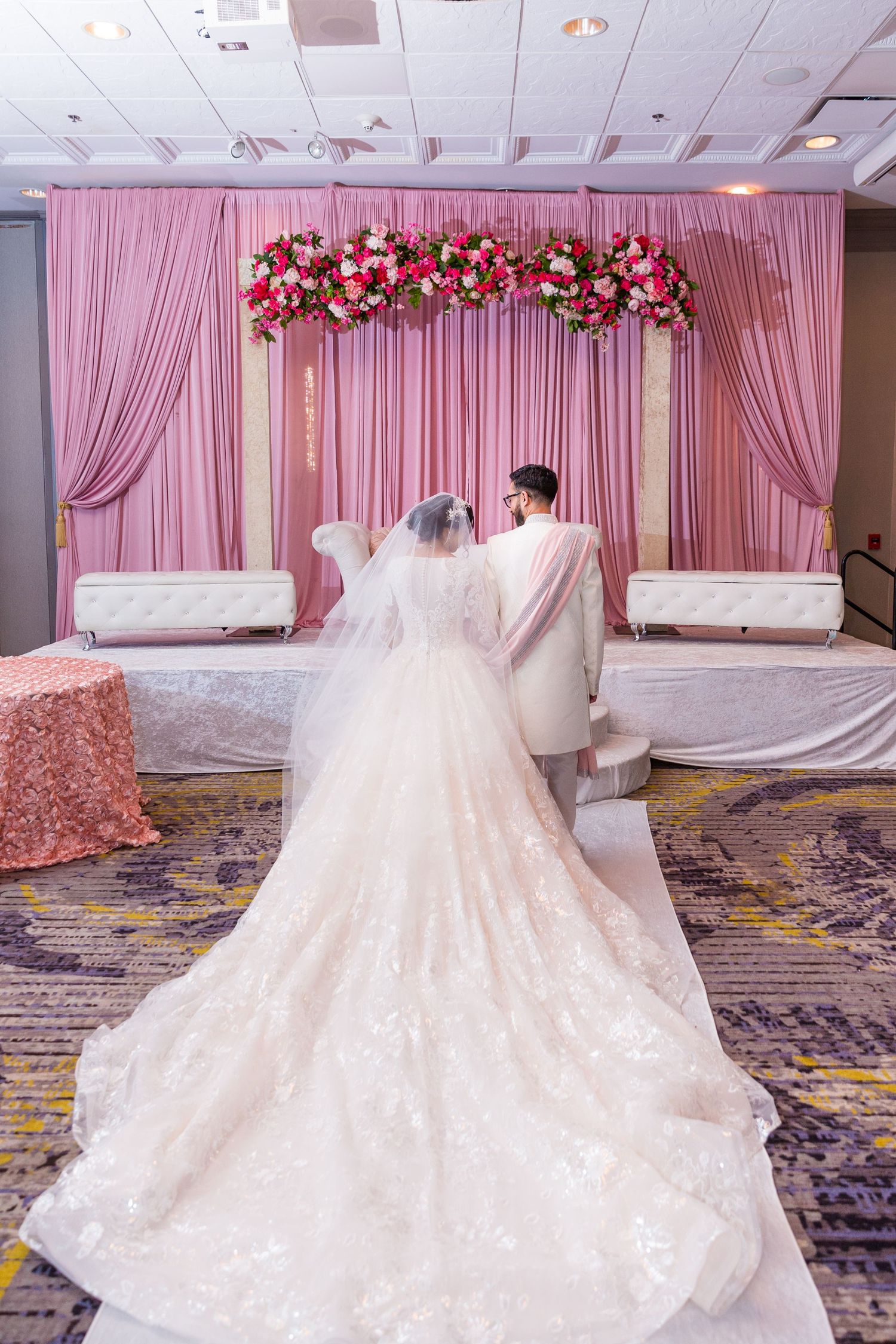 Bride and groom stand before elegant pink draped backdrop adorned with floral arrangements at wedding reception.