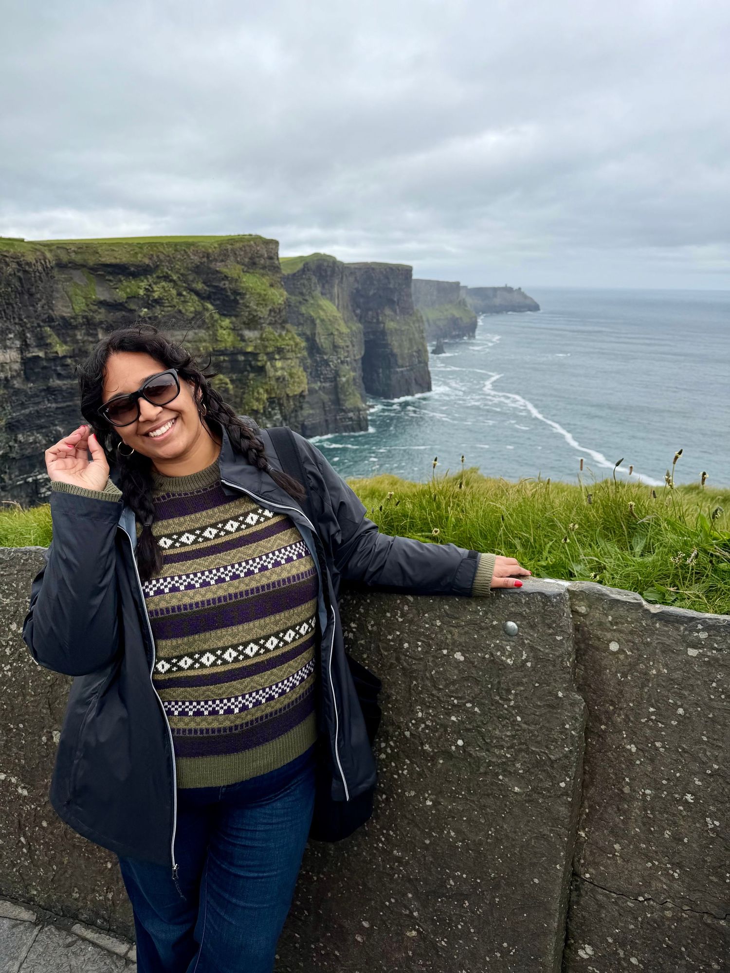 female destination wedding elopement photographer smiling at the Cliffs of Moher in Ireland wearing a patterned sweater and sunglasses.