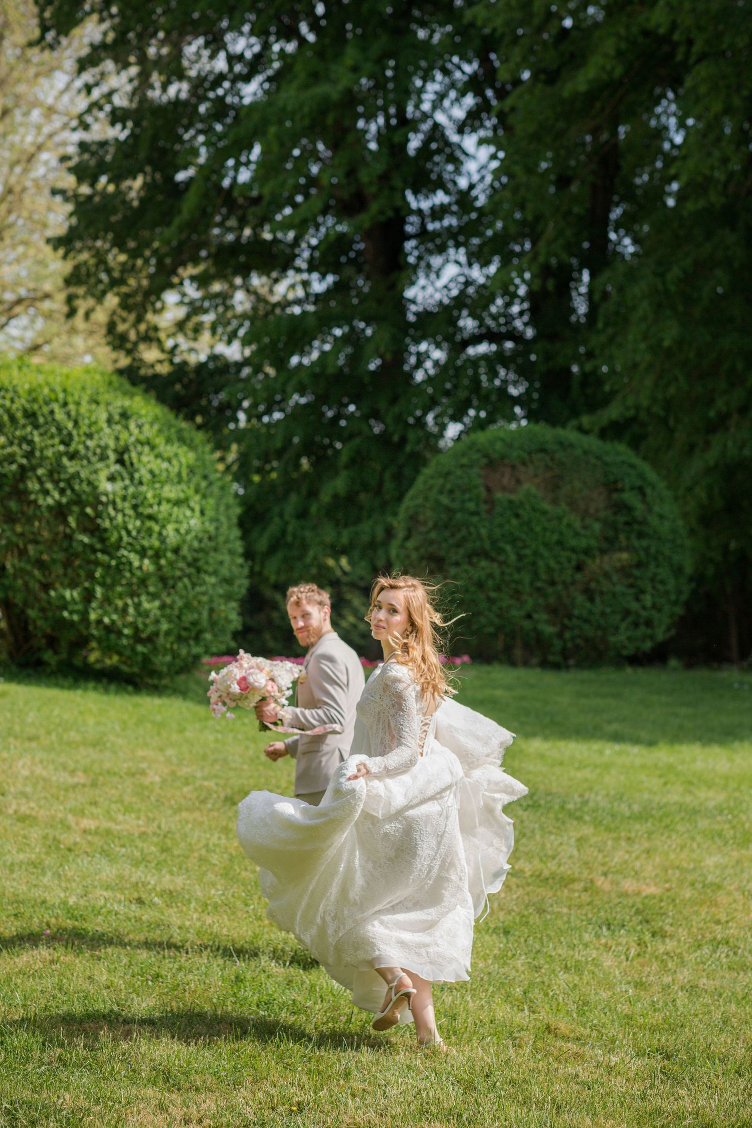 Bride twirling in white lace dress with groom holding pink bouquet in garden.