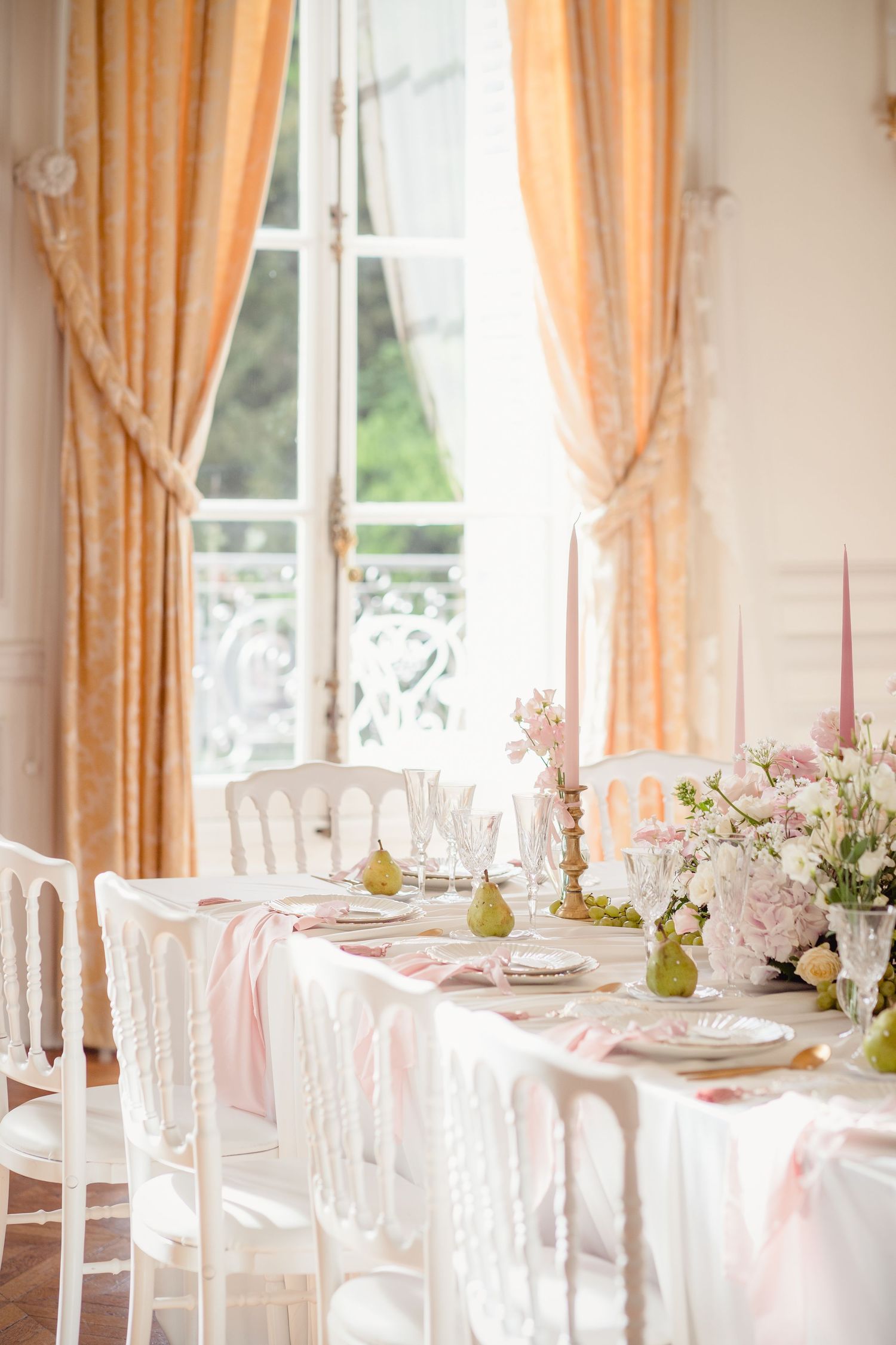Bright wedding tablescape with pink candles, pear accents, crystal glasses, and peach curtains in a French venue.