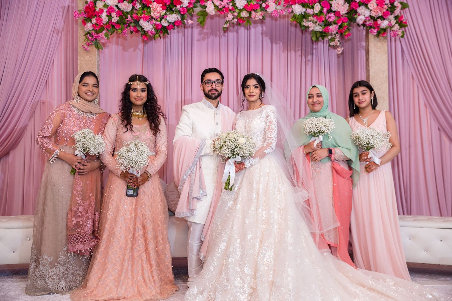 Wedding party poses in pink decor with bride in white gown, groom in white suit, and bridesmaids in pink dresses.
