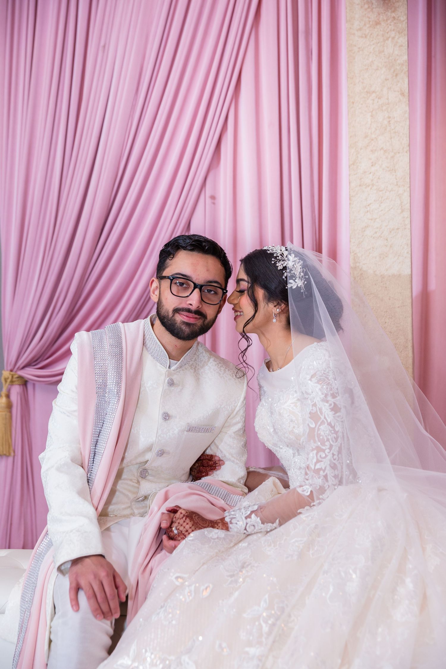 A bride and groom in elegant white wedding attire sit together against a soft pink draped backdrop.