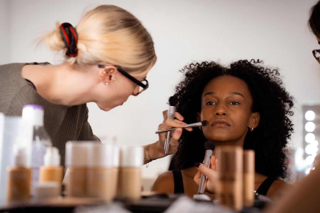 Blonde makeup artist applying foundation to young Black woman with curly hair in bright studio.