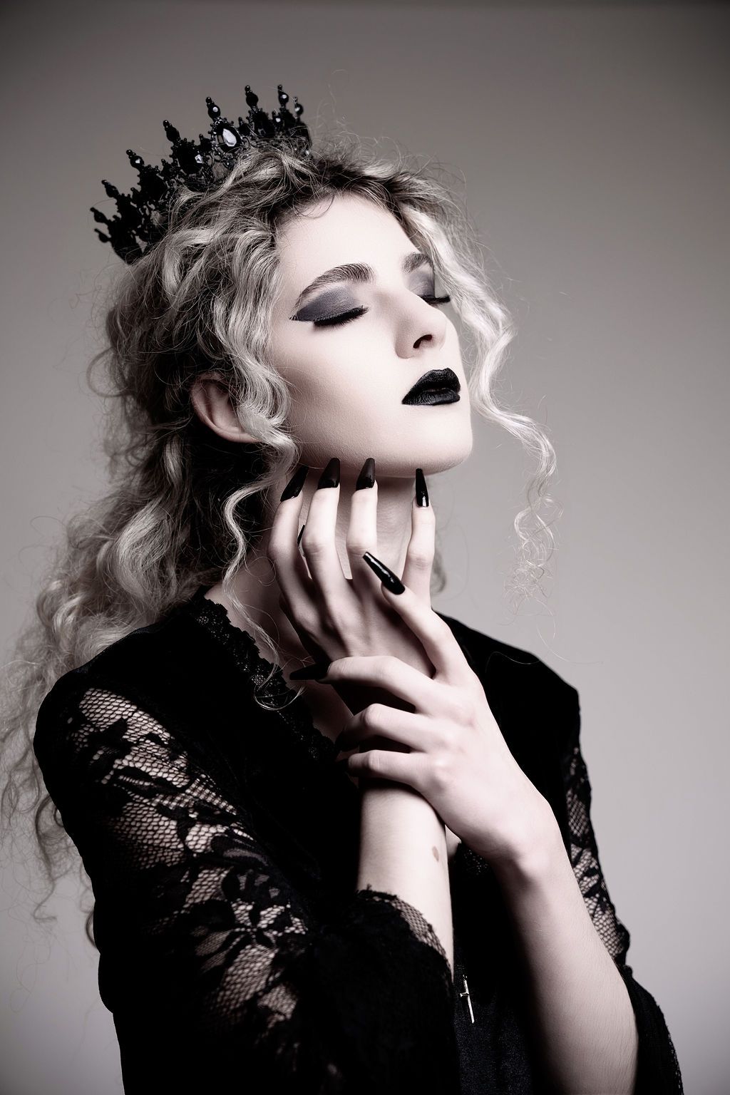 Gothic queen with black crown, dark lipstick, long black nails, and lace dress poses dramatically with eyes closed.