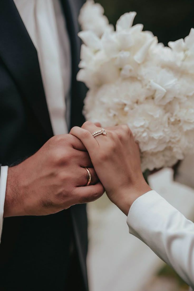Bride and groom holding hands with wedding rings and a white floral bouquet.