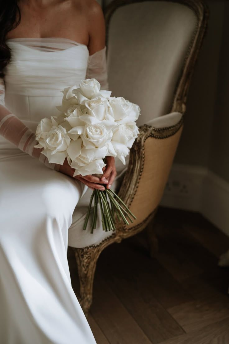 Bride in white strapless gown holding elegant bouquet of white roses while seated on vintage chair.