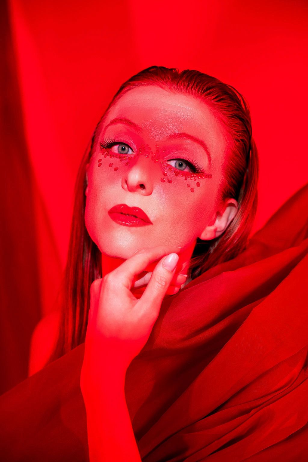 Woman with red monochromatic makeup and rhinestones posing against a red background with flowing fabric.