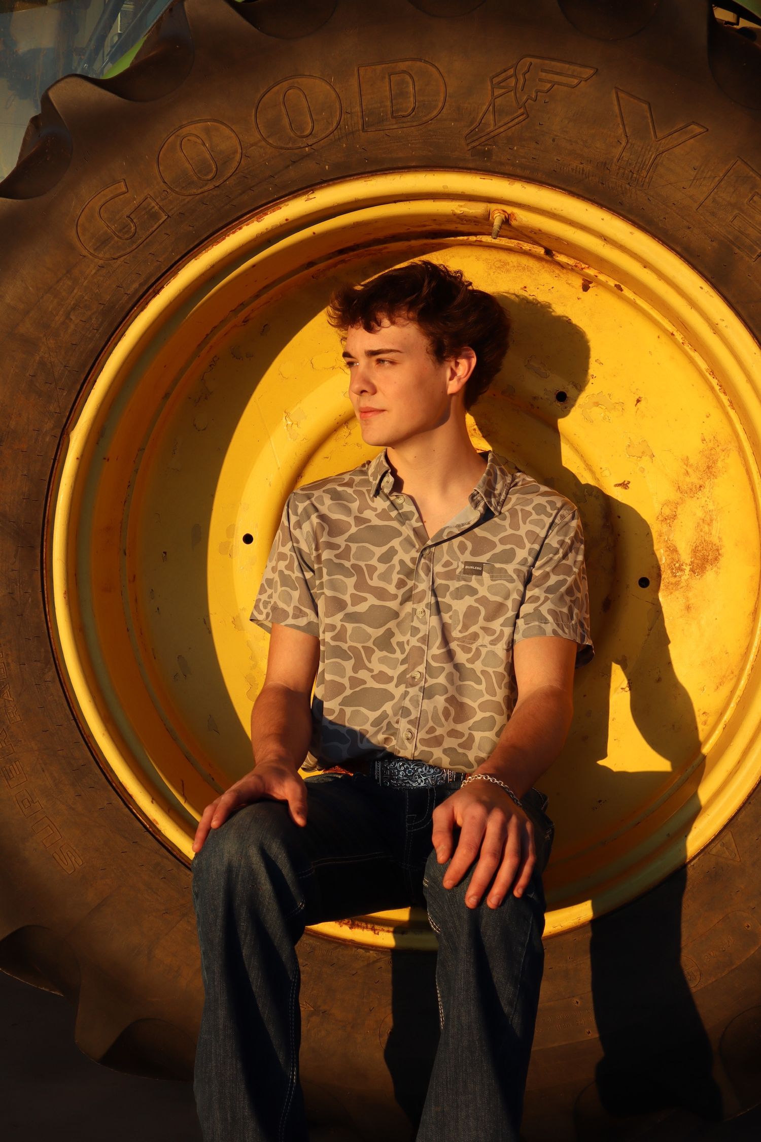 Person sits against large yellow tractor tire at sunset wearing camouflage pattern shirt and jeans.