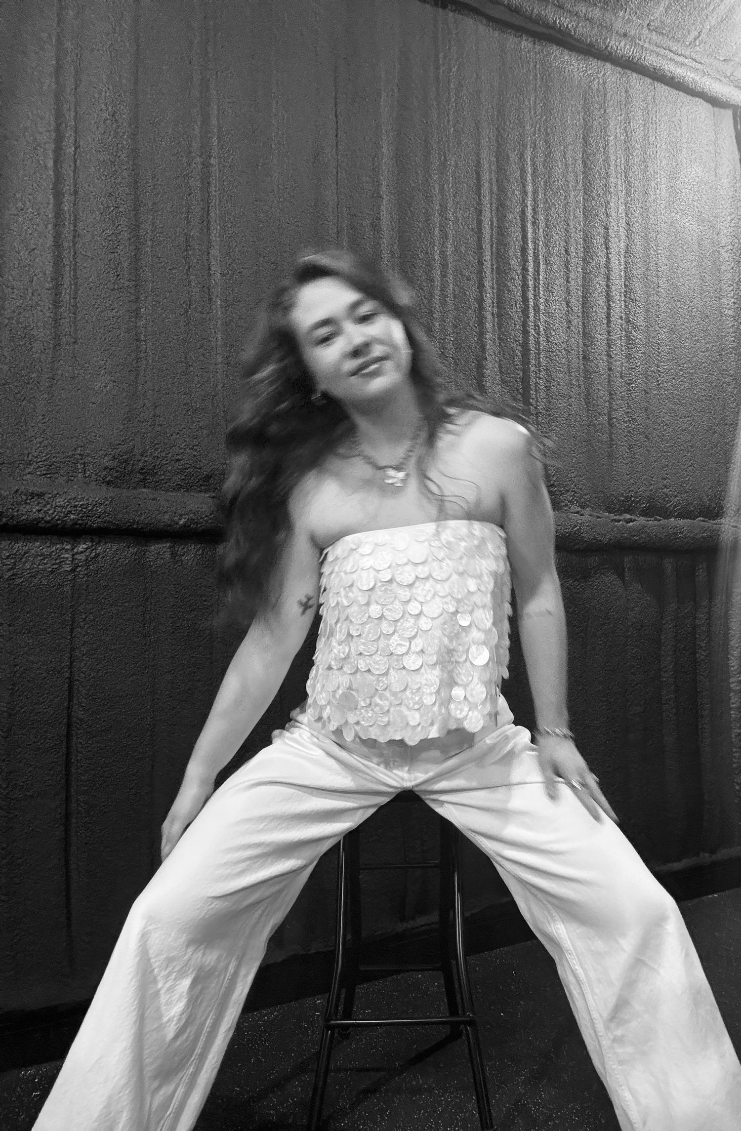 A black and white portrait of someone in a white lace top and pants sitting casually on a stool.