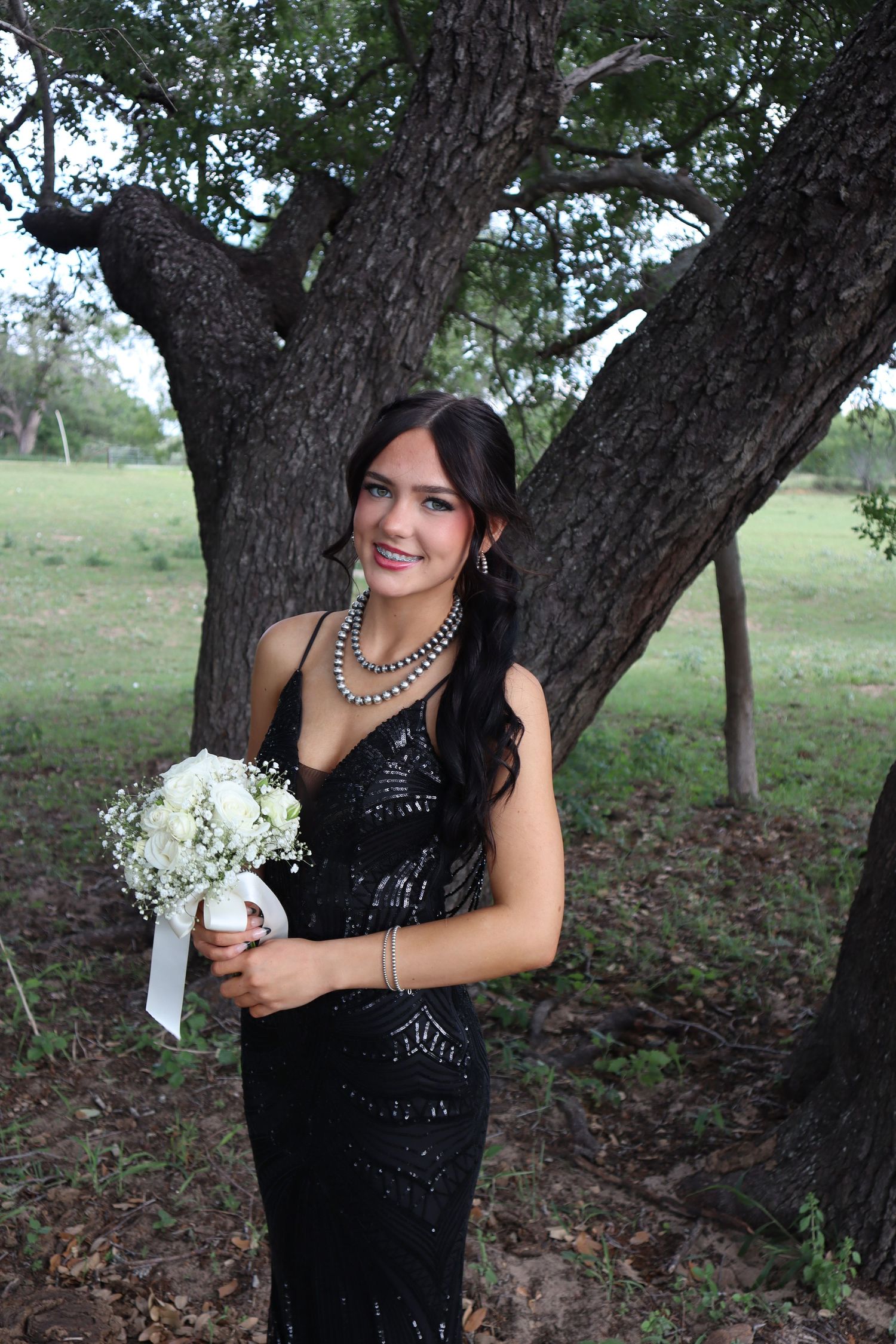 A formal evening photo shows someone in a black sequined gown holding a white bouquet near a large tree.