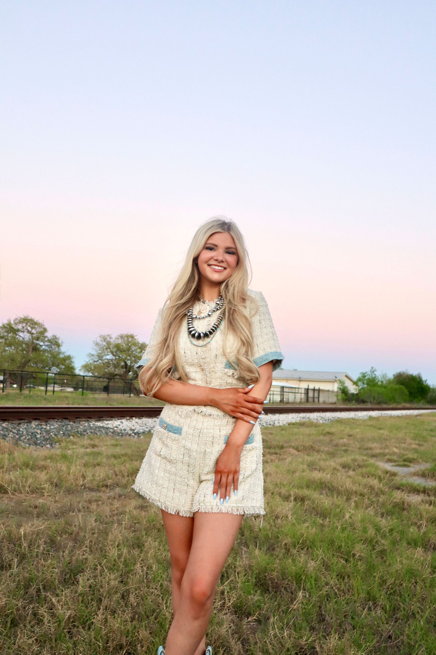 A person in a white lace dress and statement necklace poses outdoors at sunset against a rural backdrop with railroad tracks.