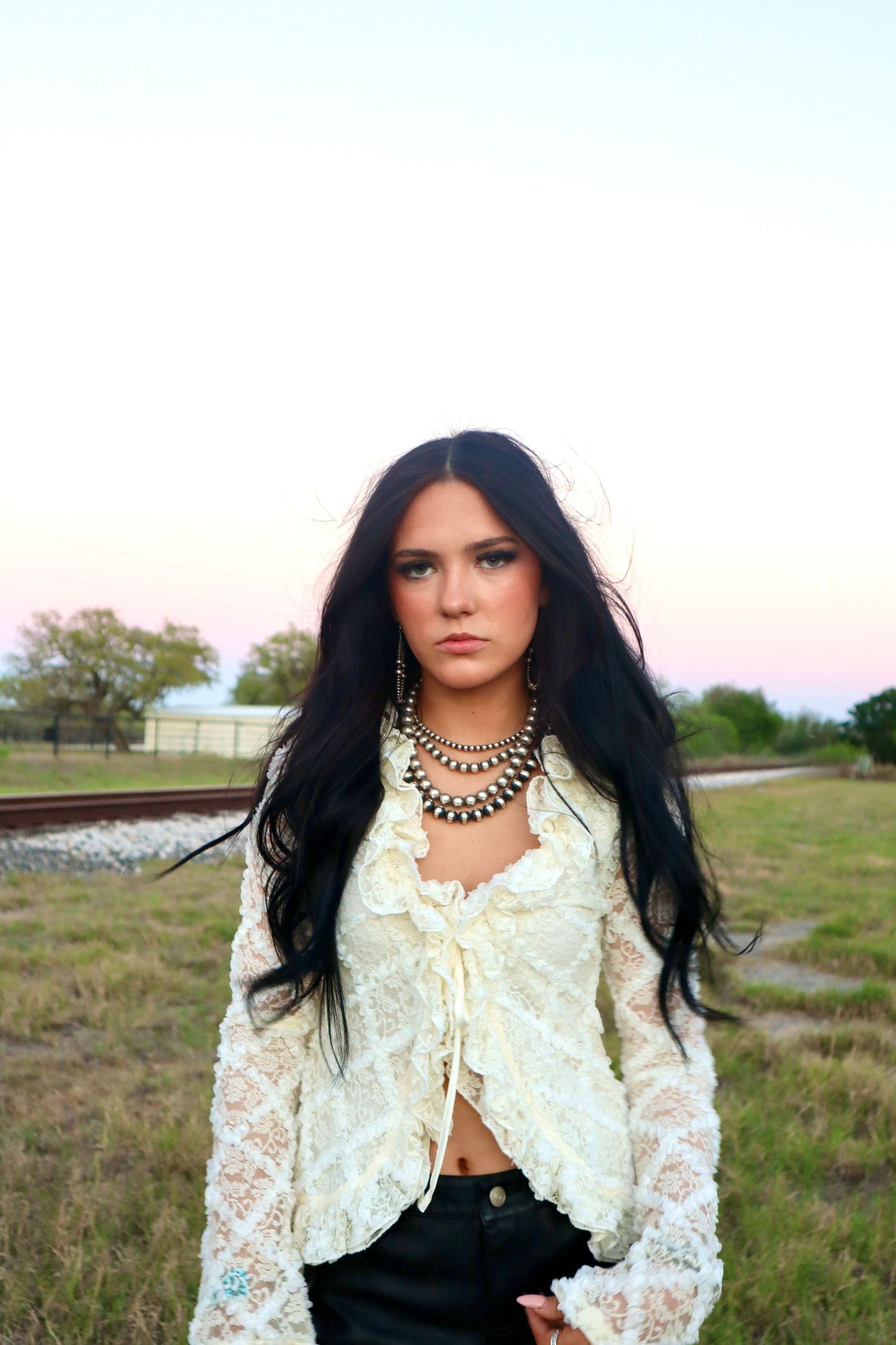 A woman with long dark hair poses in a white lace top and black skirt in a rural outdoor setting at sunset.