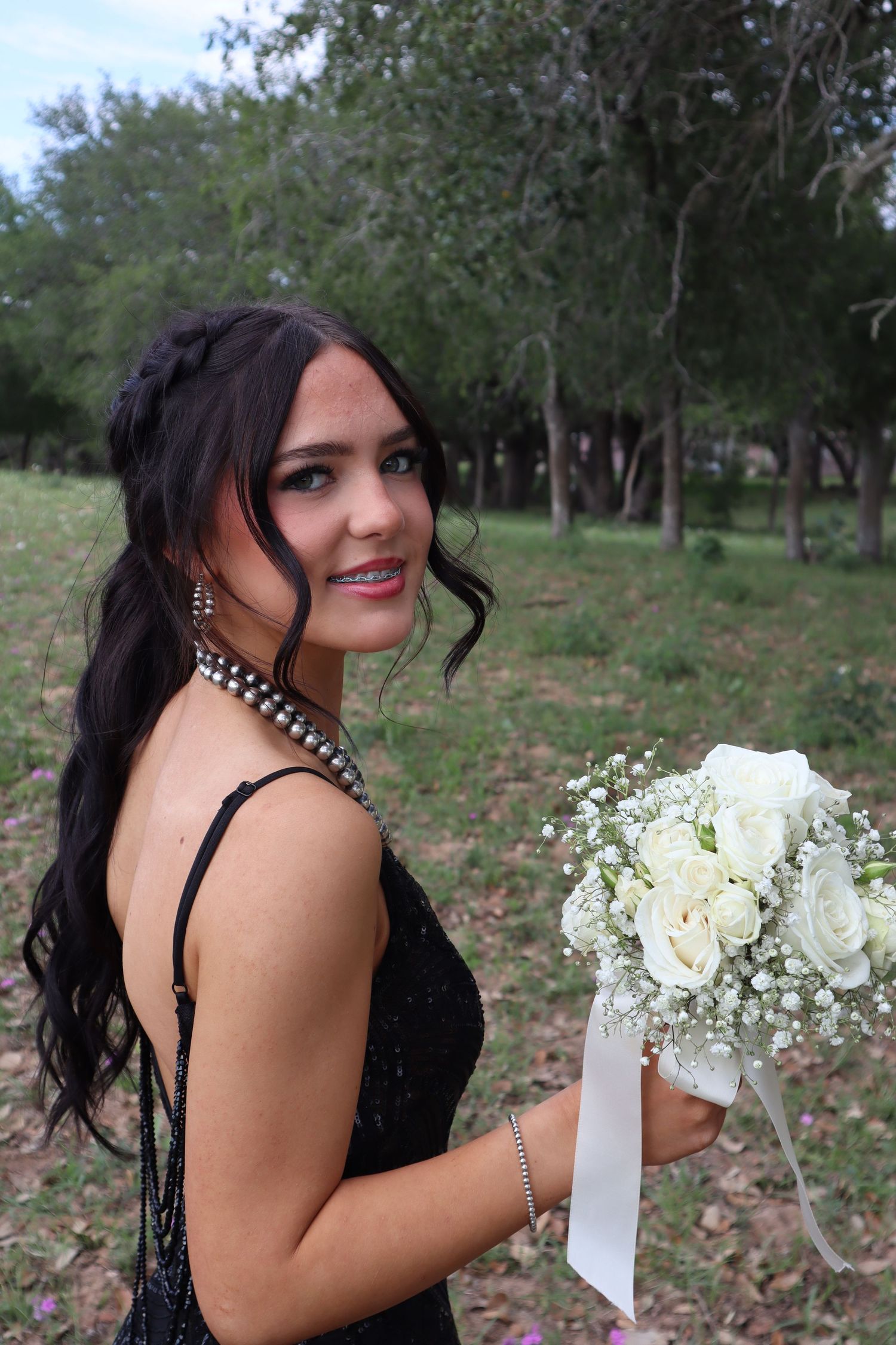 Someone in an elegant black dress holds a white floral bouquet while posing outdoors among trees in a natural setting.