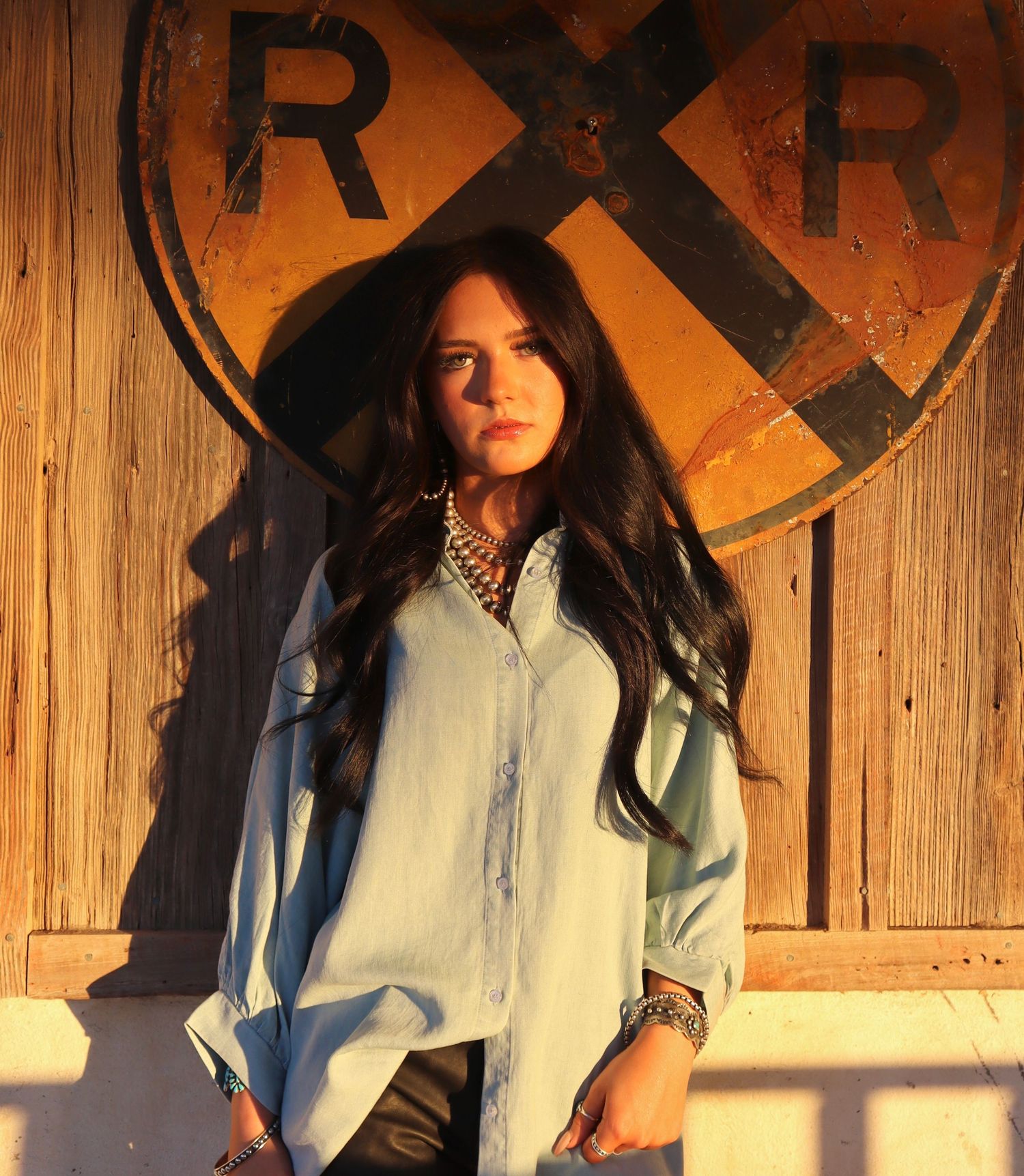A person in a denim jacket stands against a wooden wall with railroad crossing signs in warm lighting.